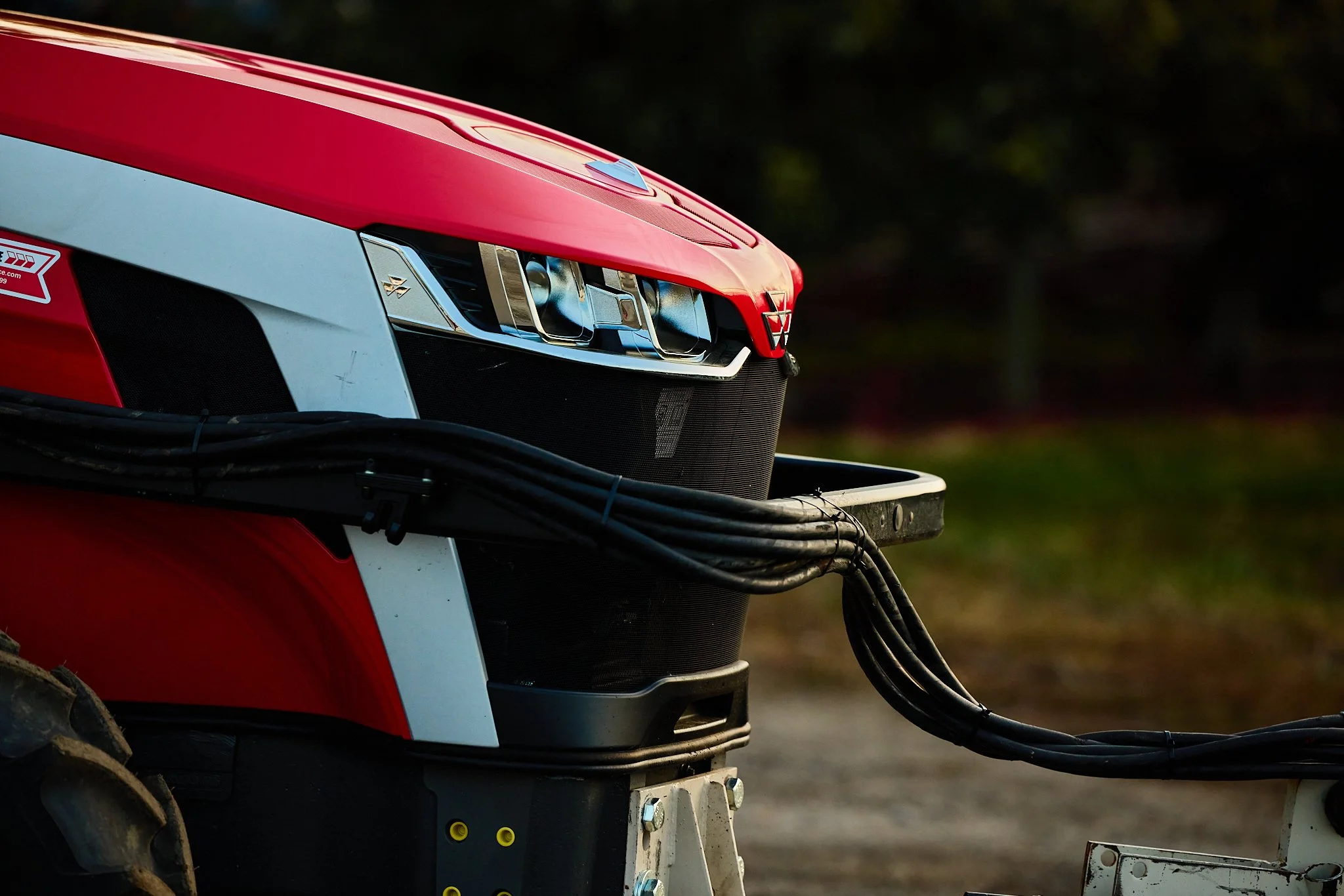 Close-up of a modern red tractor's front, showing its headlights, grille, and black wiring harnesses, with a blurred outdoor background.