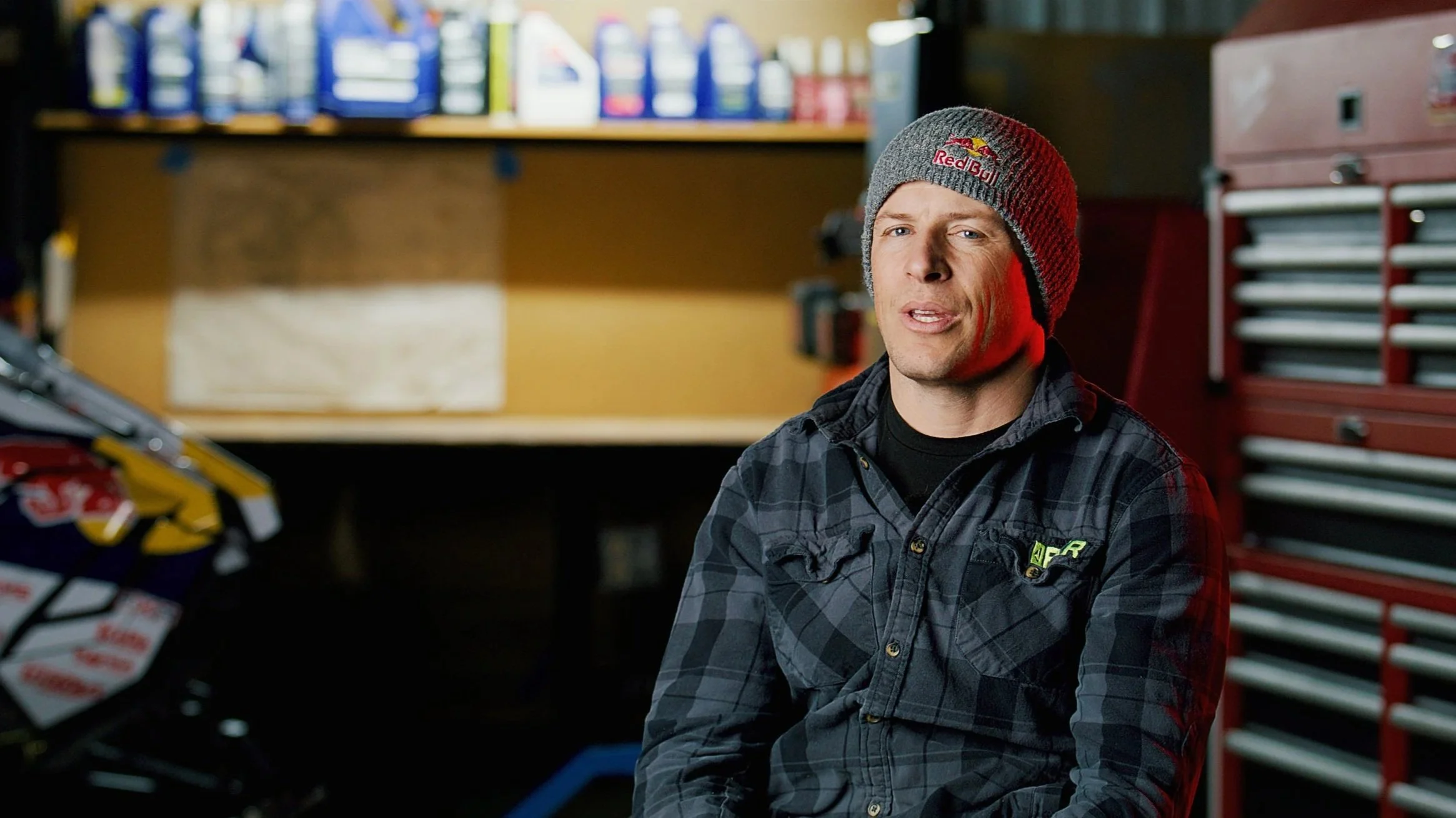 A man wearing a Red Bull beanie and a black checkered shirt sitting in a garage with a motorcycle to his left and red toolboxes behind him.