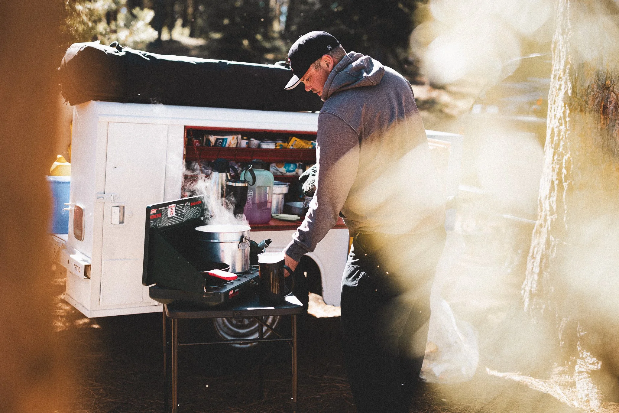 A man wearing a gray hoodie and black cap preparing food on a portable stove outdoors near a white trailer.