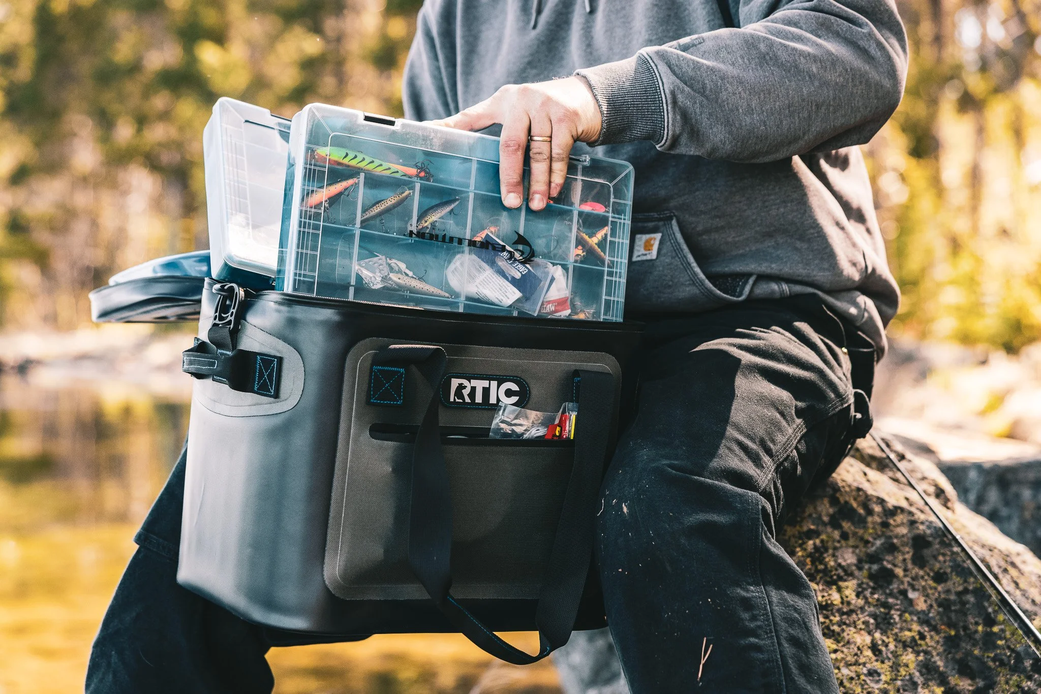 Person sitting outdoors on a rock, organizing fishing lures and tackle in a clear plastic storage box, with a fishing rod nearby, during autumn.