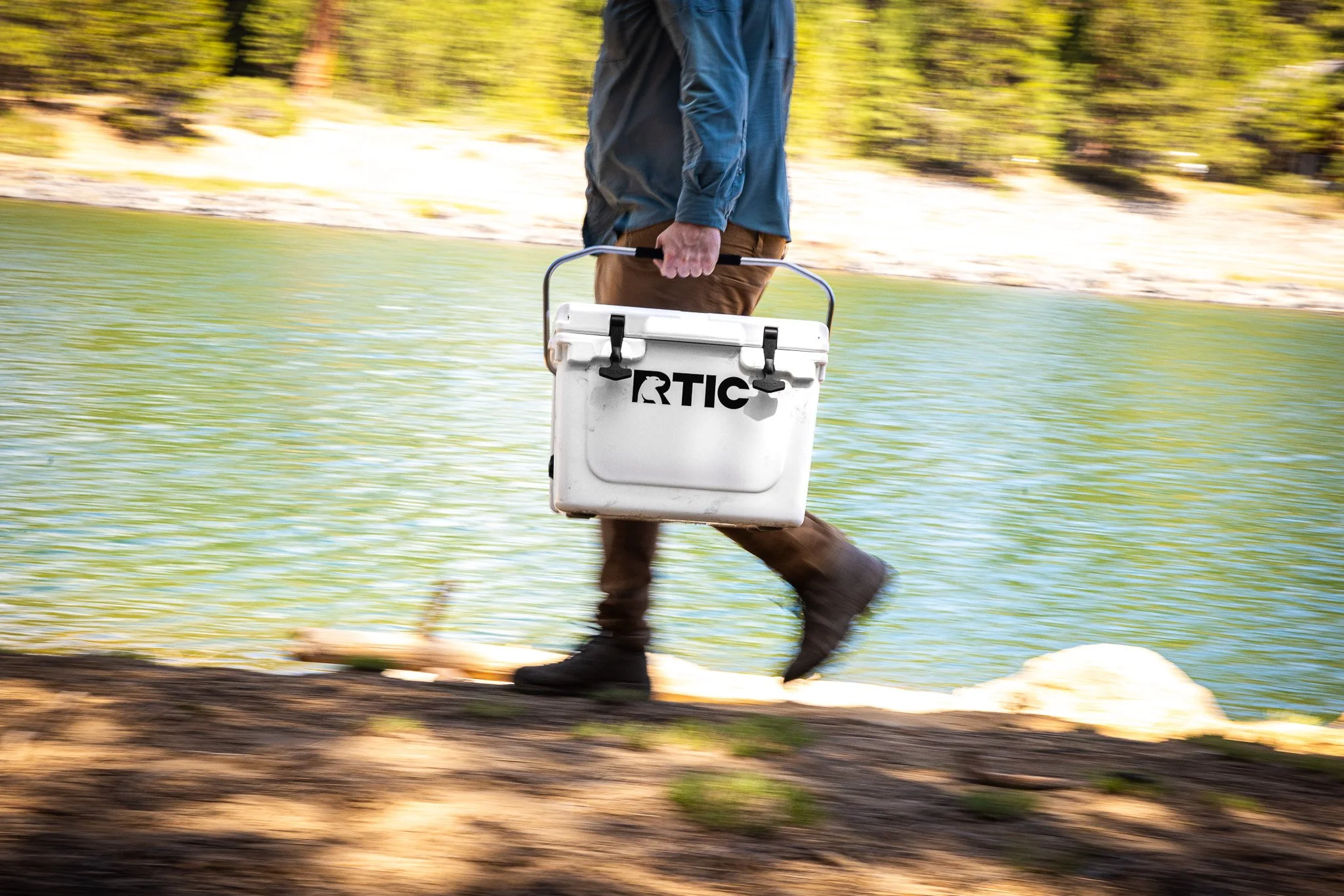 Person walking along a lakeshore carrying a white cooler.