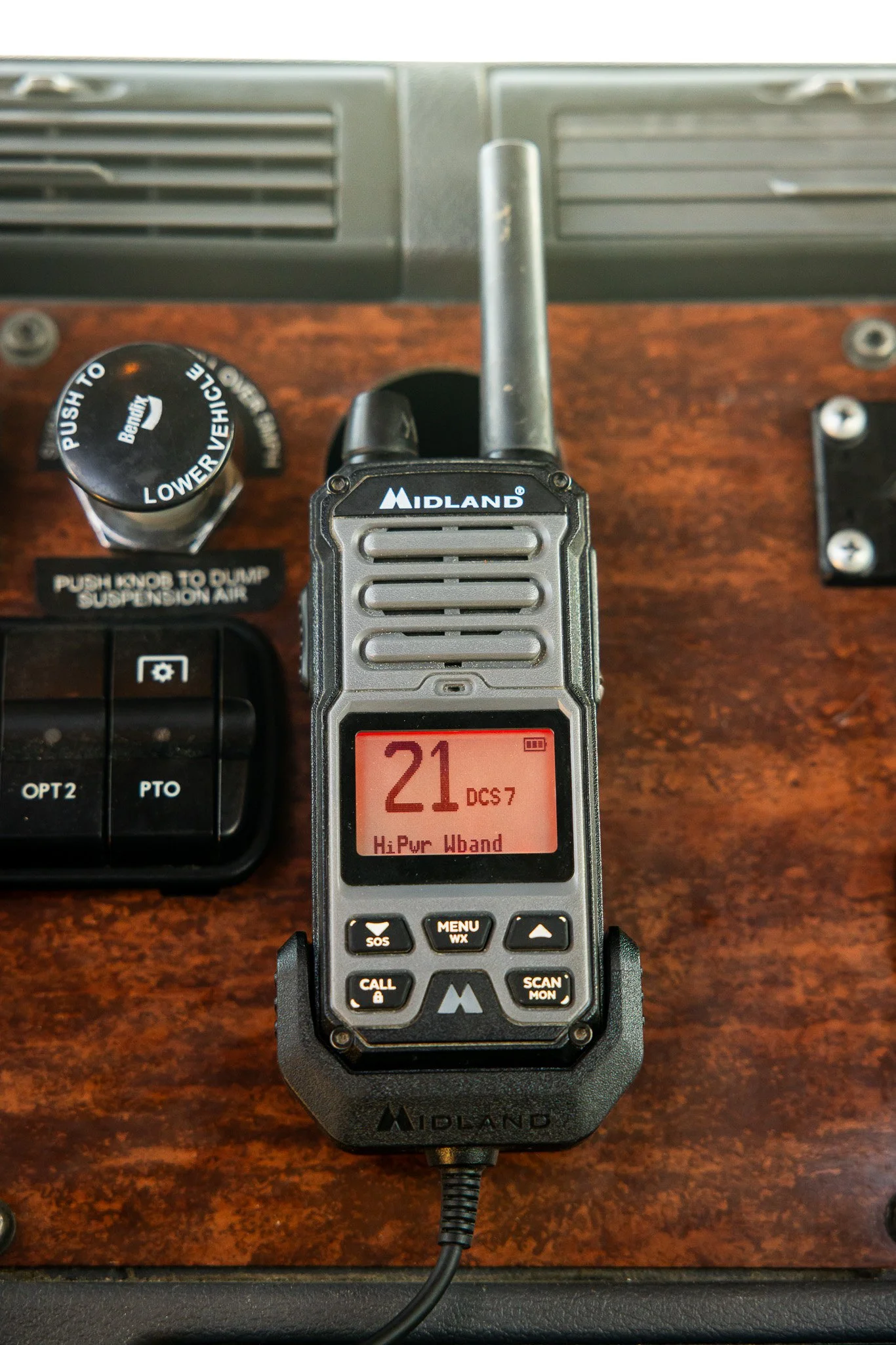 A Midland two-way radio with a red display showing the number 21, connected to a charging base on a wooden surface. Other radio controls and a vent are visible in the background.