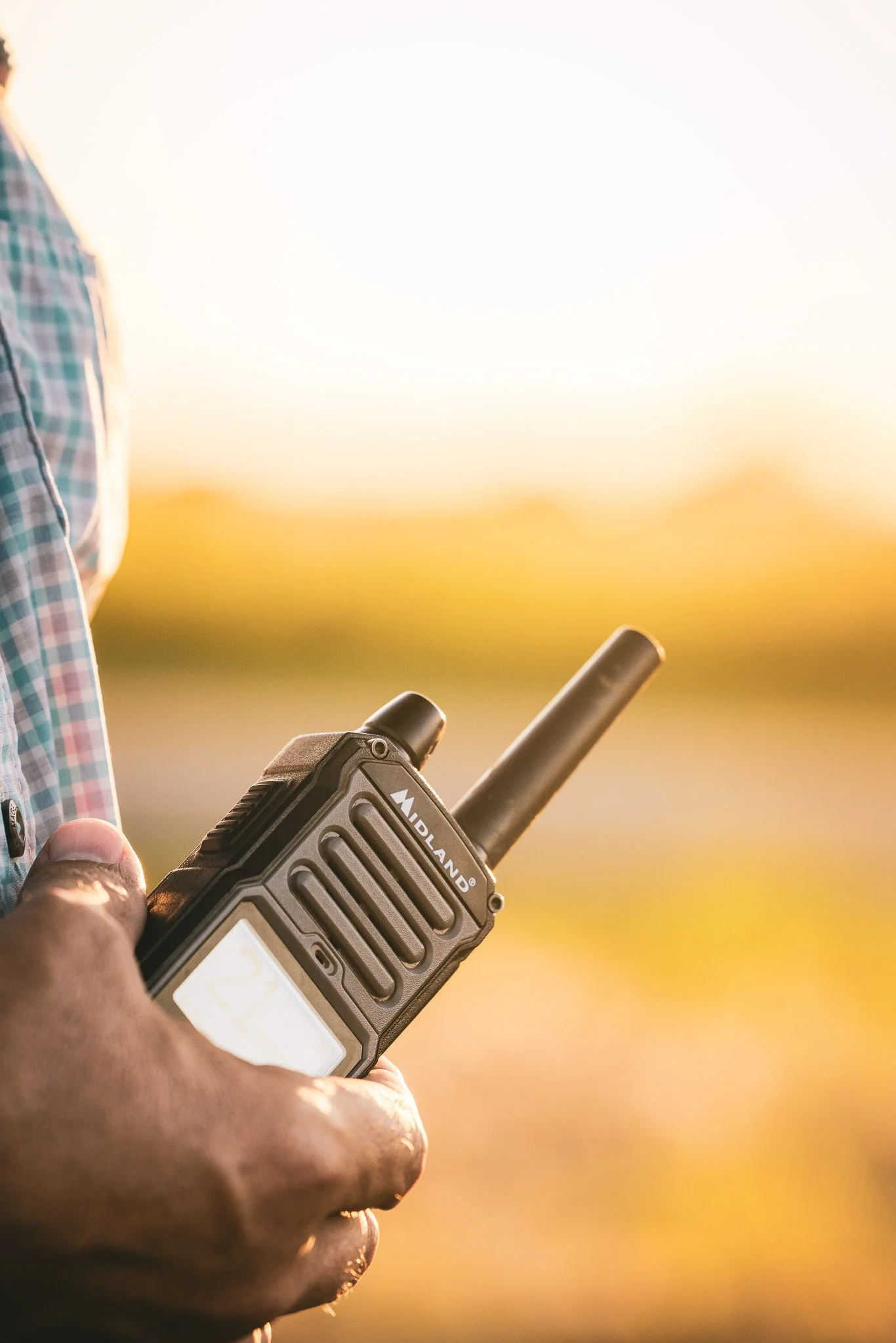 Close-up of a person's hand holding a Midland walkie-talkie with a sunset or sunrise in the background