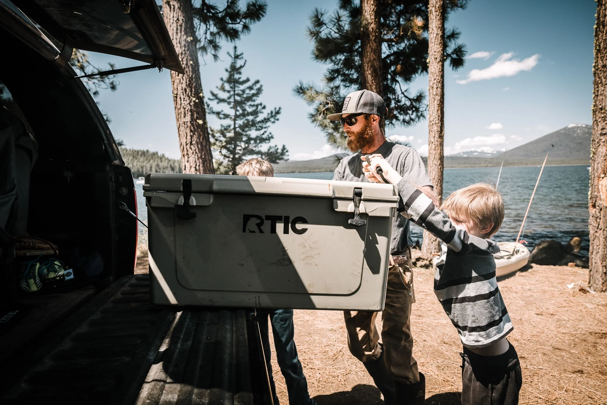 A man and two children by a lake, with a kayak and a cooler, surrounded by pine trees and mountains under a blue sky.