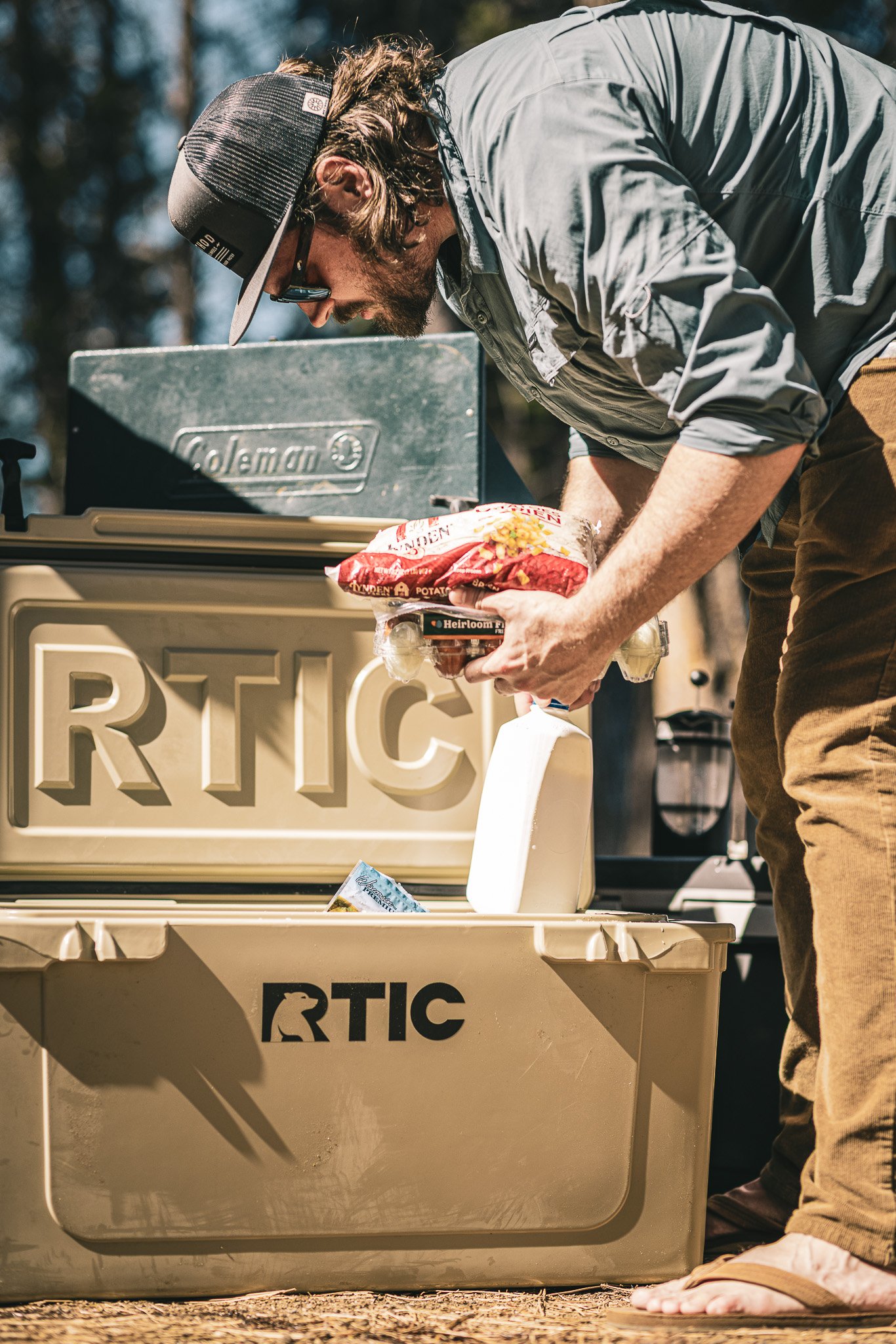 A man wearing a gray shirt, brown pants, a black cap, and glasses is leaning over a beige RTIC cooler outdoors on a sunny day, packing it with food items, including a bag of potatoes and a container of Heirloom tomatoes.