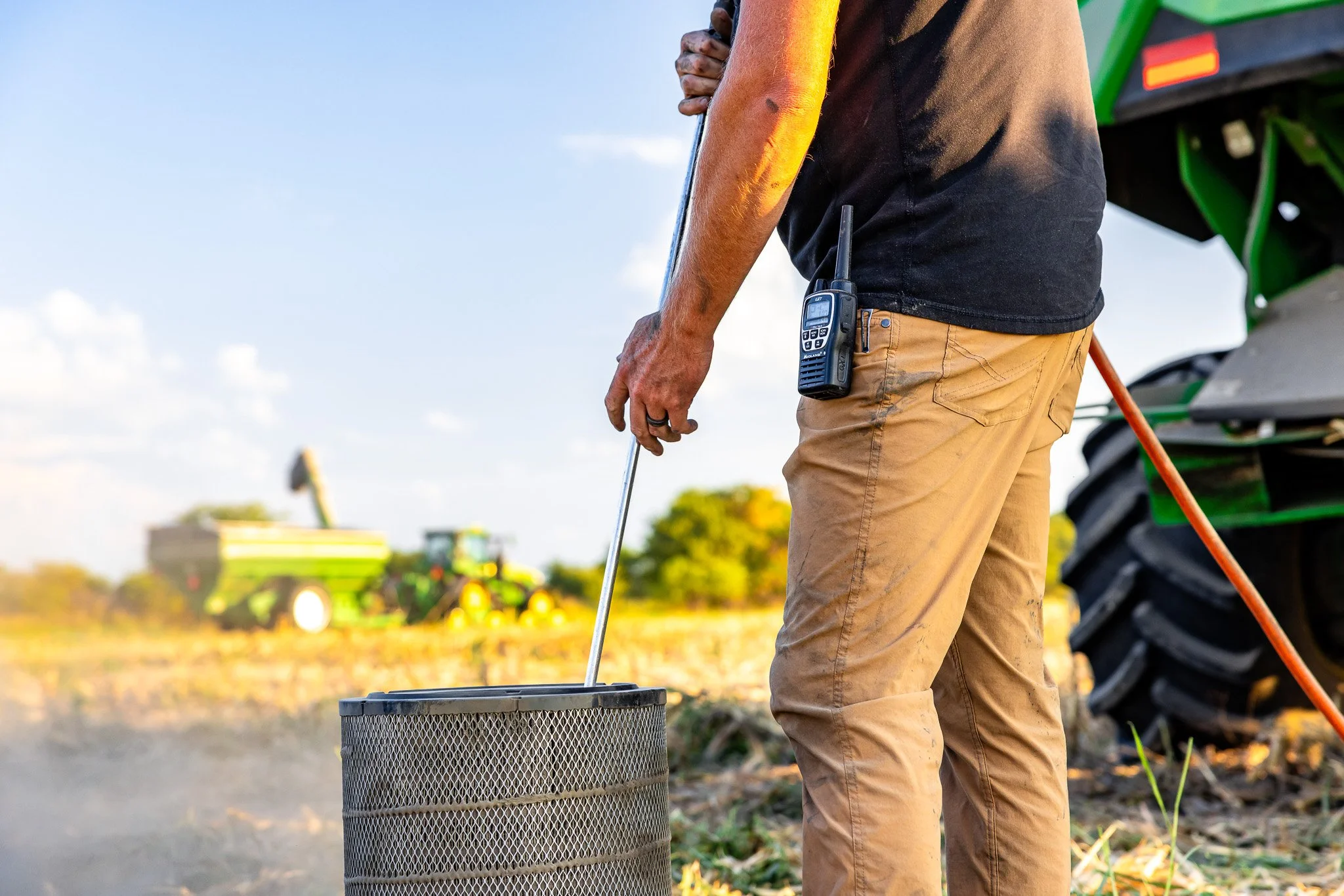 Farmer standing outside on farmland, using a metal probe for soil testing, with a walkie-talkie clipped to his black t-shirt, and a green tractor with large tires in the background.