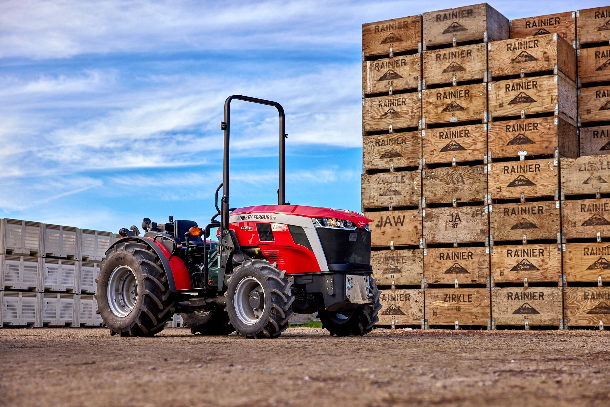 Red Massey Ferguson tractor parked on dirt in front of stacked wooden crates with labels 'Rainier' and others, under a partly cloudy sky.