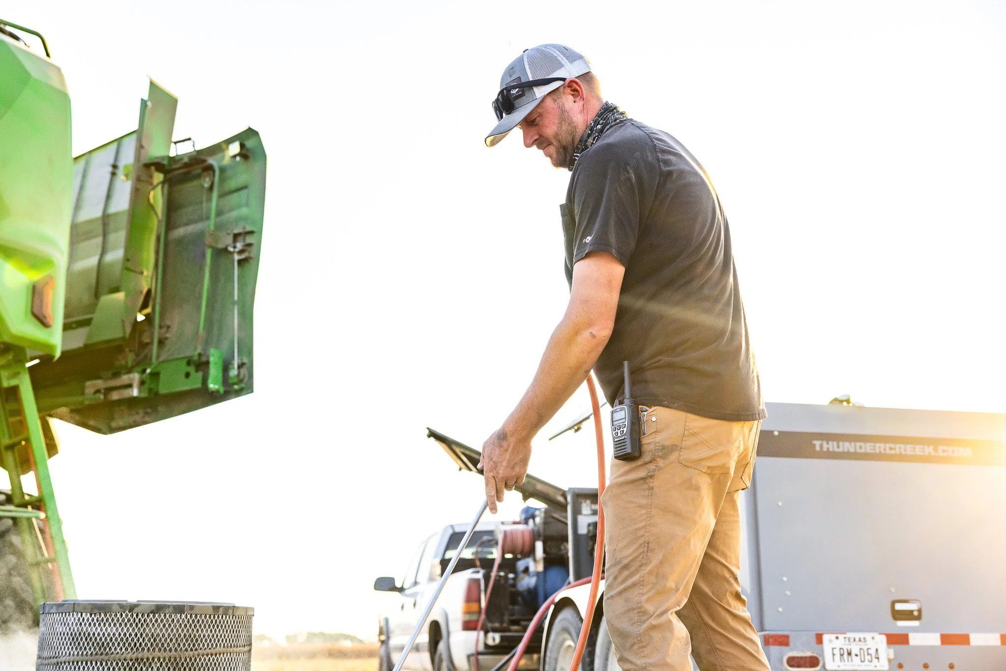 A man wearing a black shirt, beige pants, sunglasses, and a hat is working outdoors on electronic equipment connected to a green machine, with a pickup truck and a trailer in the background.