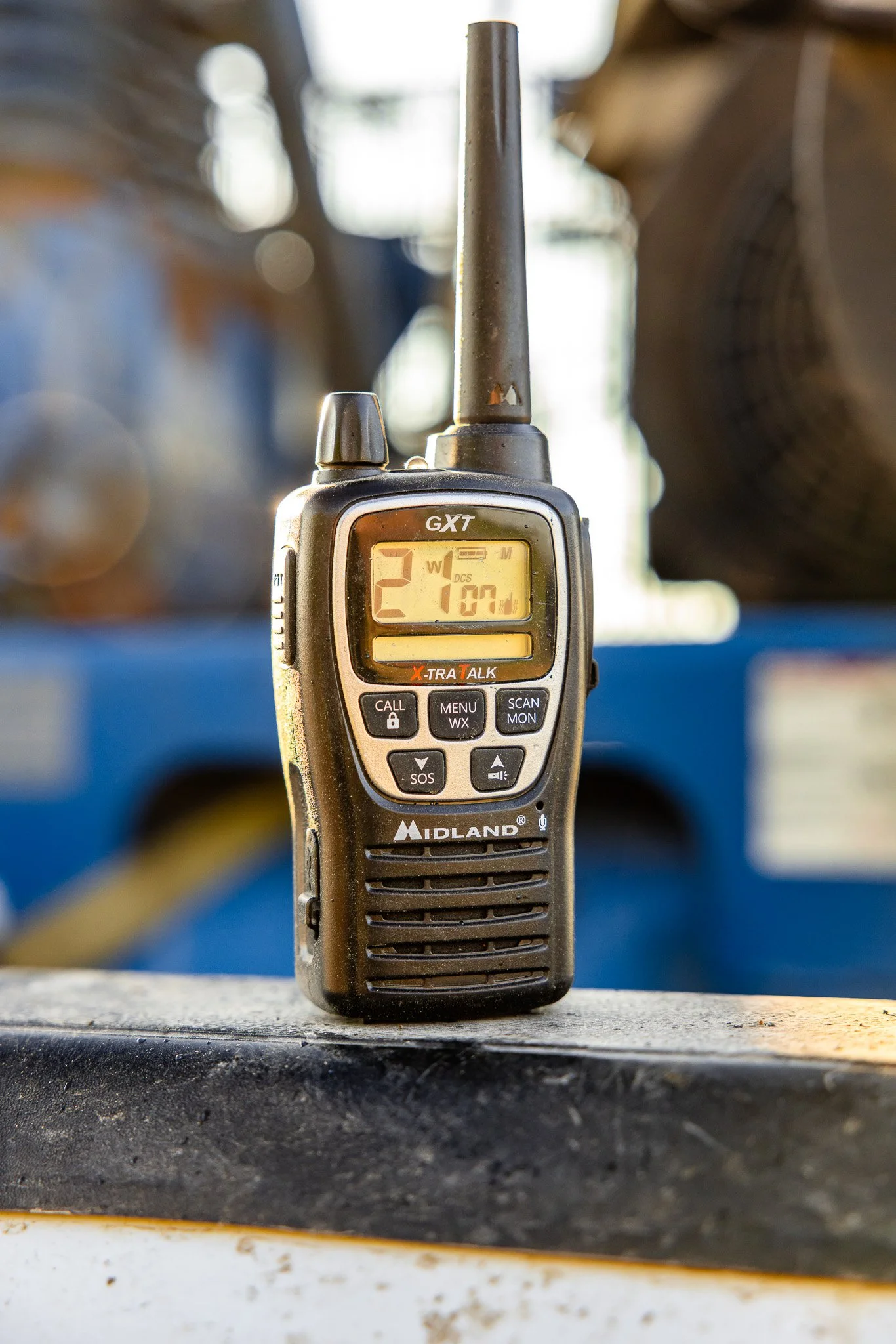 A Midland GXT handheld walkie-talkie with a digital display showing 21.7. It is placed on a surface in a workshop with blurred tools and equipment in the background.