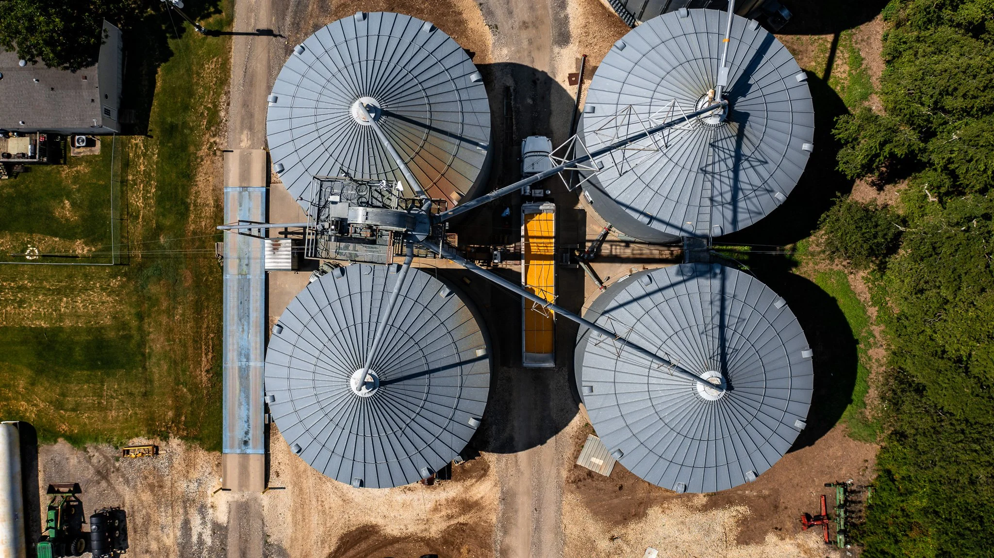 Aerial view of four large grain silos with metal roofs, connected by conveyor systems, surrounded by green trees and a dirt area with machinery.