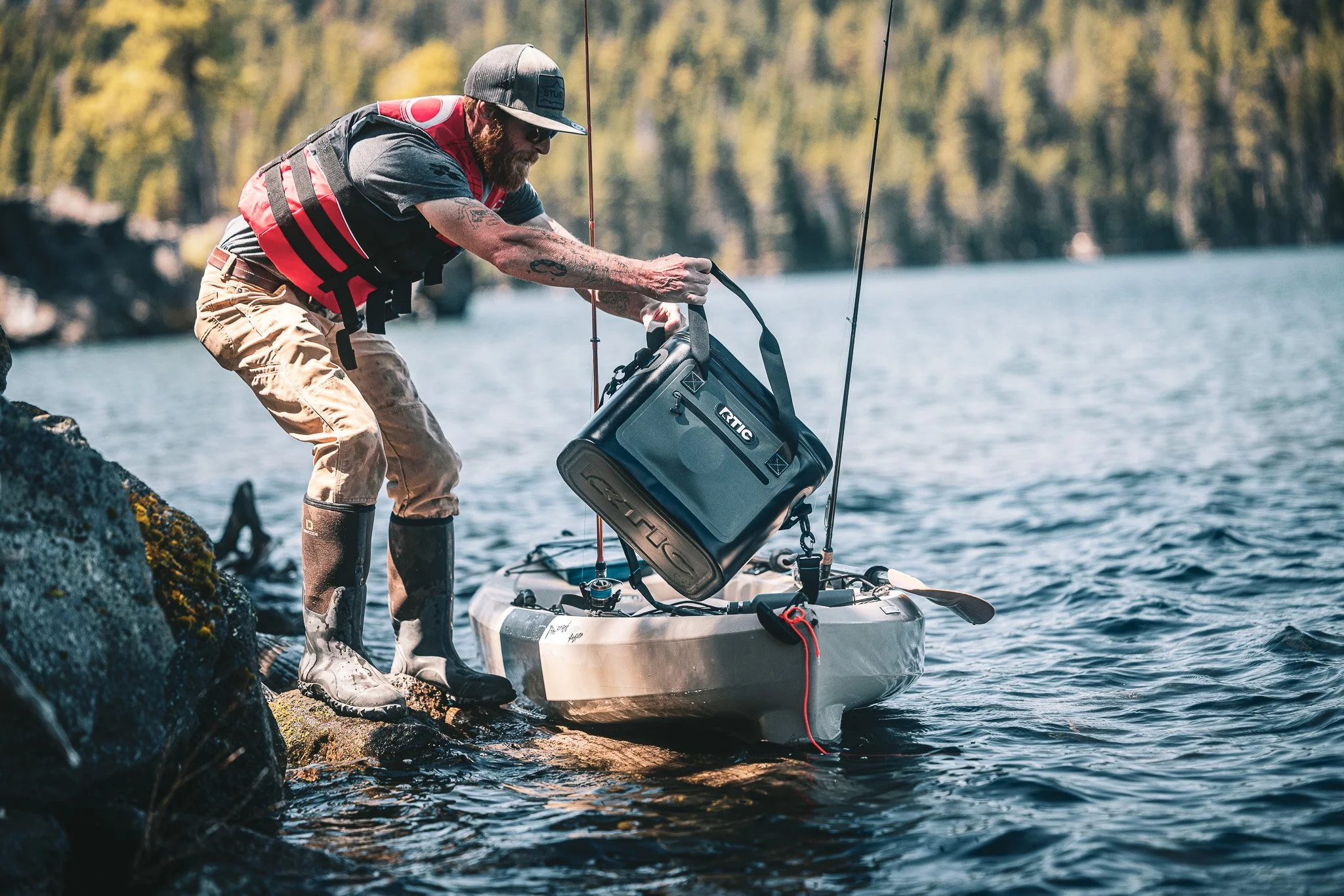 A man with a beard, wearing a gray cap, sunglasses, and waterproof boots, is leaning over a small boat or kayak on a rocky shoreline, placing a gray portable speakers into the boat. The man is dressed in a red life jacket, a gray t-shirt, and camoufl