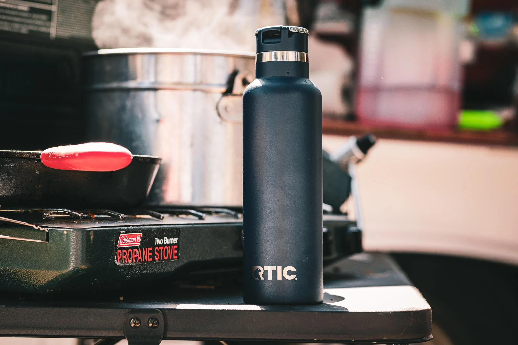 A black RTIC water bottle placed on a small outdoor stove with a red lid resting on a pan. In the background, there is a large metal pot and various outdoor kitchen items.