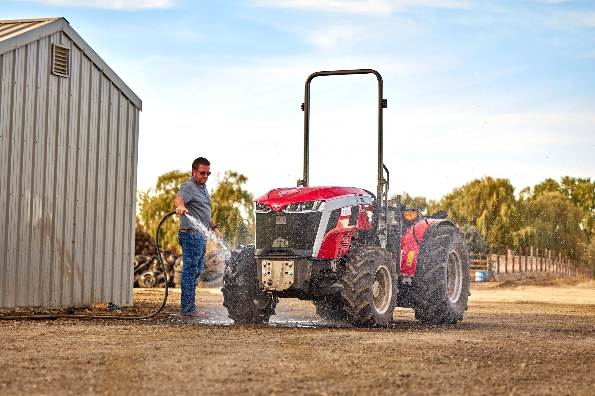Man washing a red tractor with a hose outside near a building and trees.