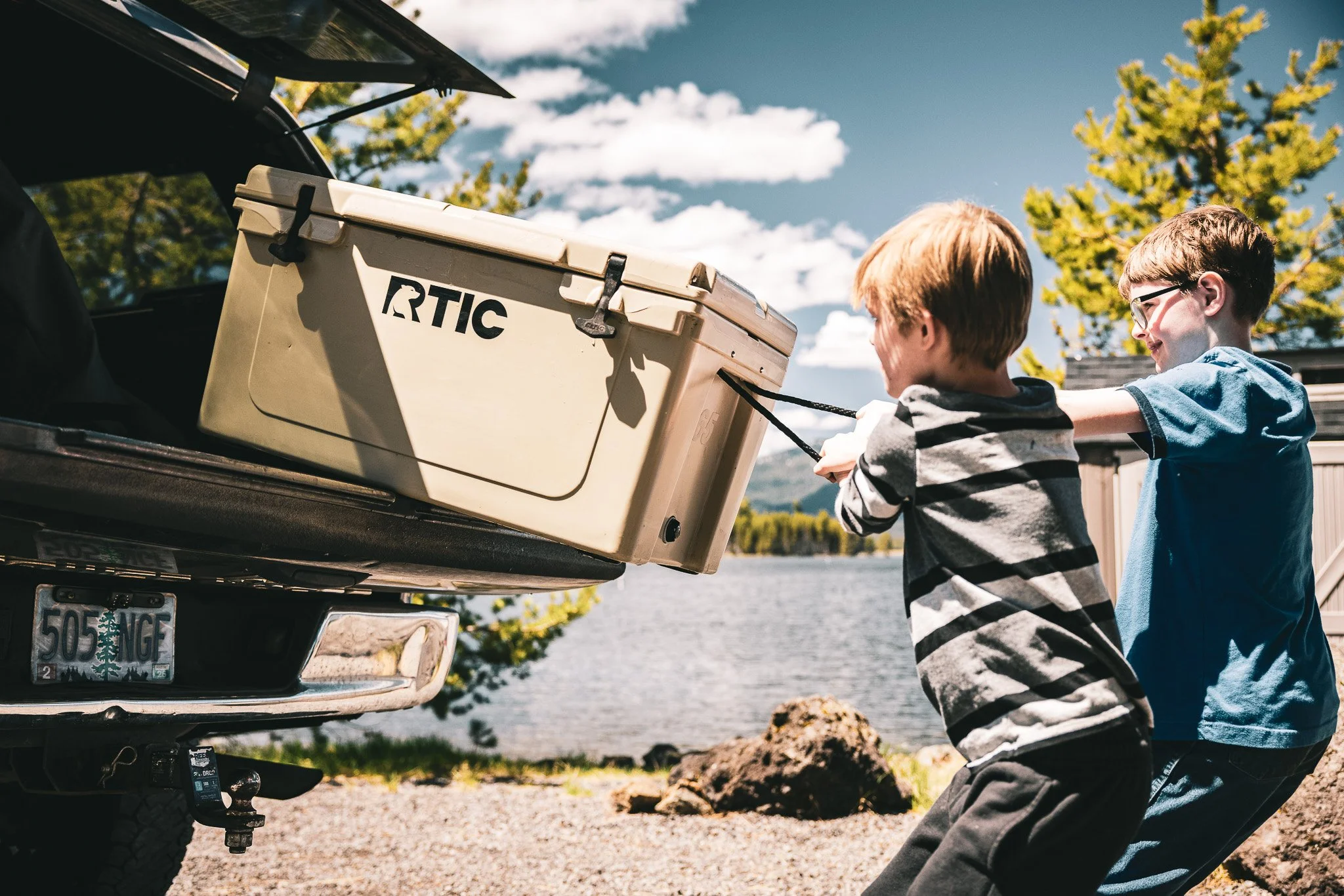 Two boys pulling a cooler out of the trunk of a car near a lake, with trees and a cloudy sky in the background.