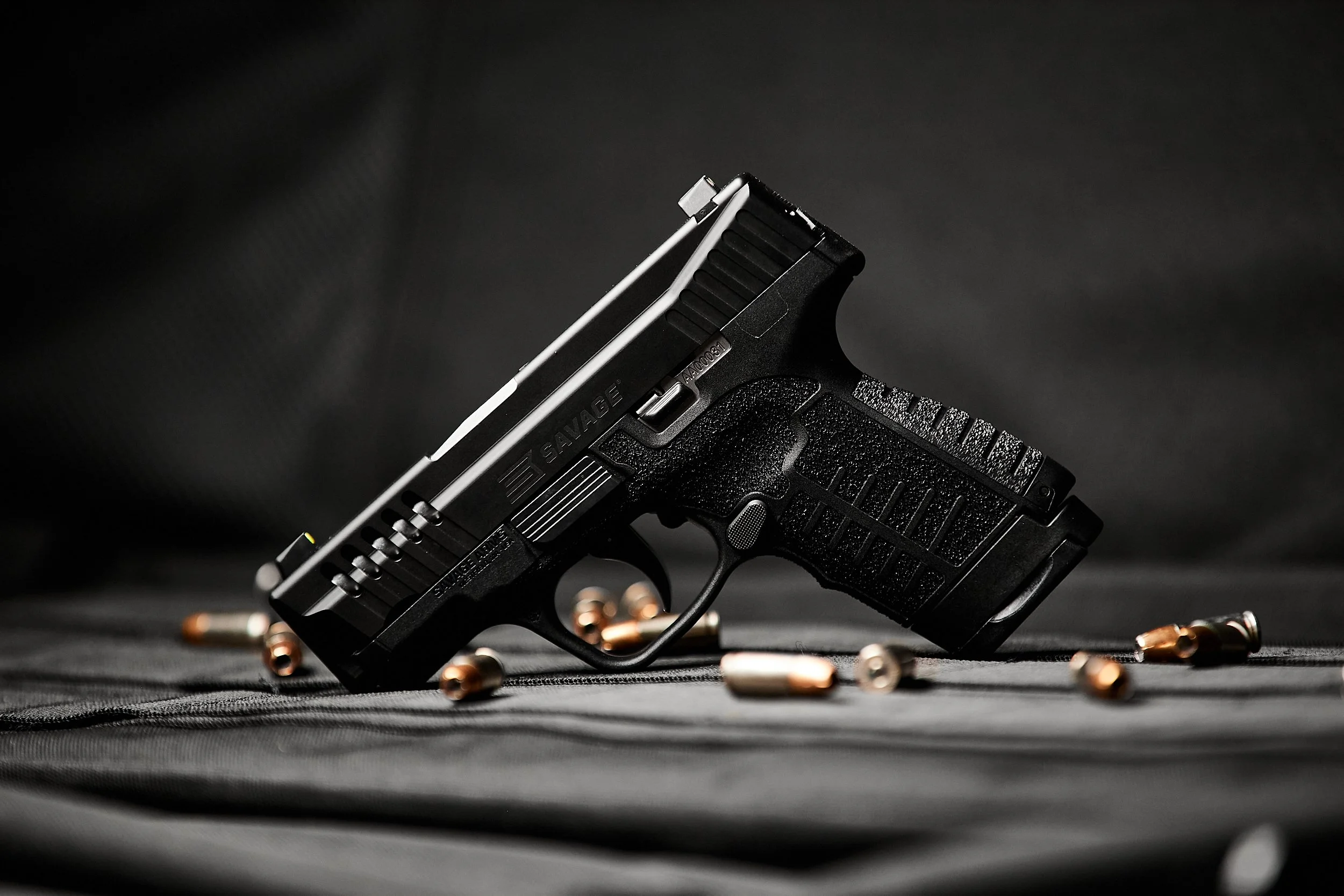 A black semi-automatic pistol with ammunition scattered around it, resting on a dark fabric surface with a dark background.