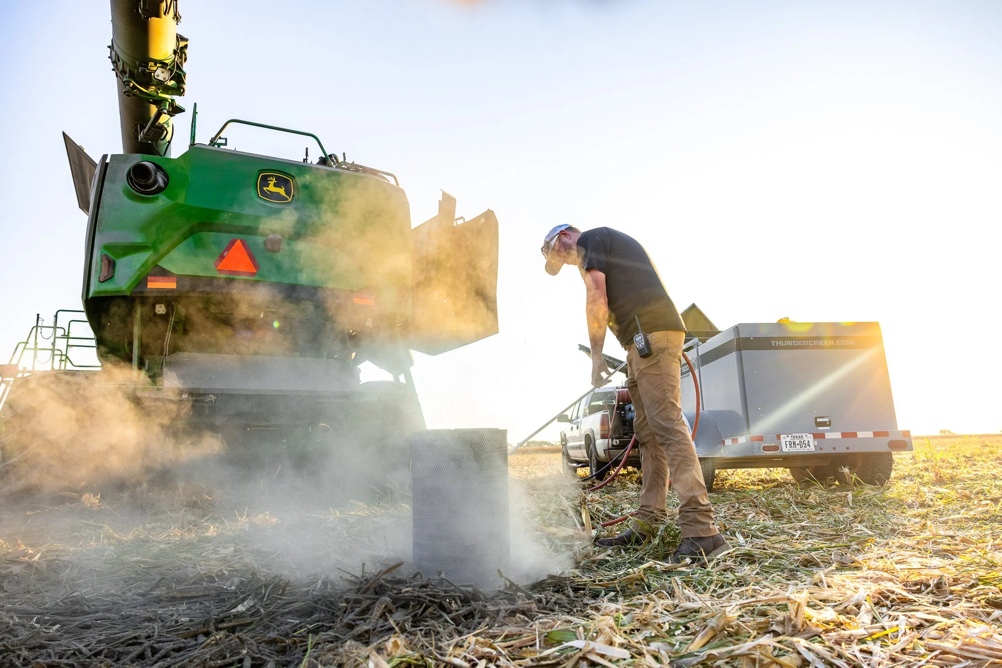 A person operating a piece of farm machinery in a field with dry crop remnants, with a trailer attached behind him, during sunset or early evening.