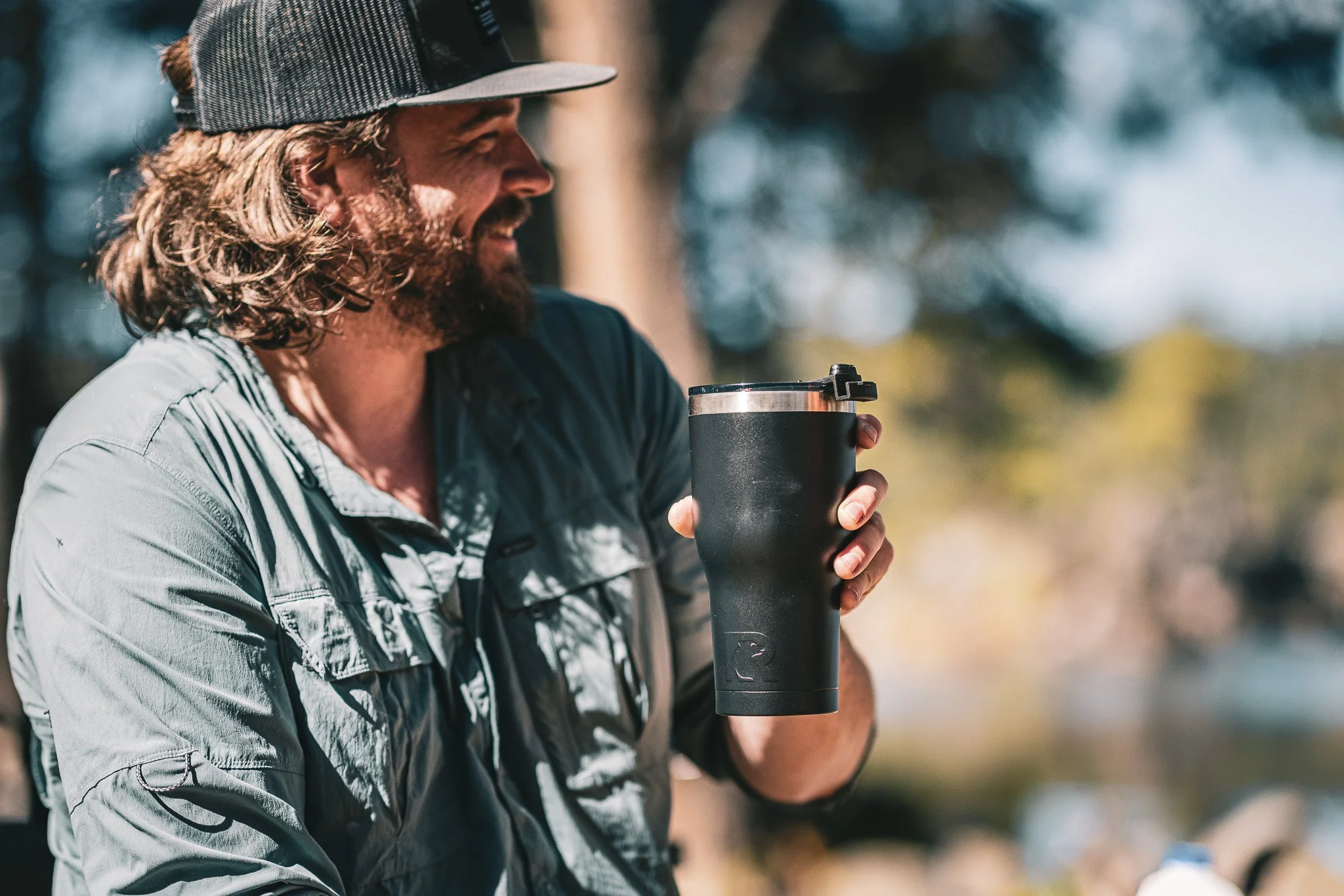 Man with long hair, beard, wearing a cap, smiling, holding a black travel mug outdoors with trees in the background.