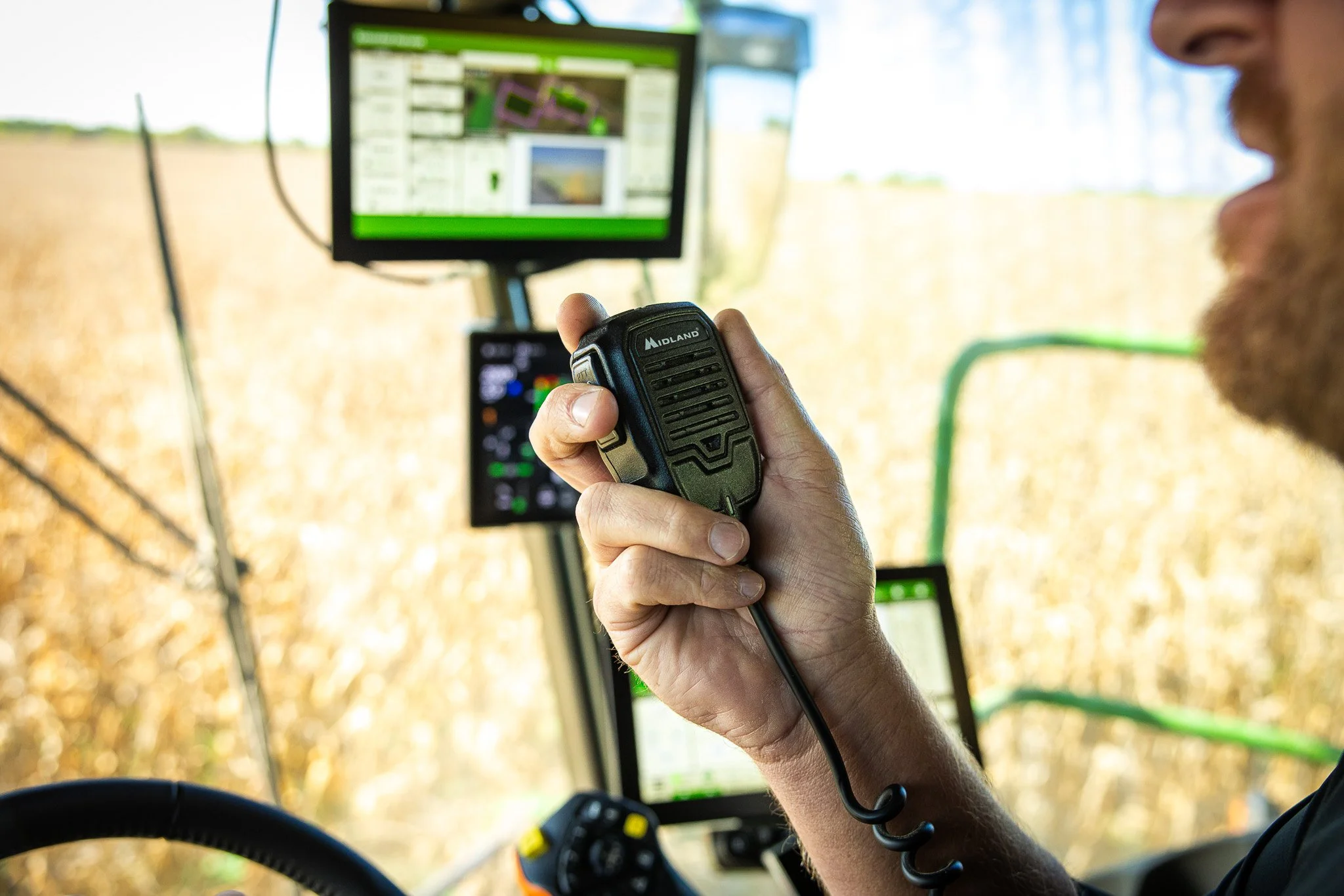 Farmer operating a tractor using a radio or communication device, with multiple screens displaying agricultural data on a bright, sunny day in a field.