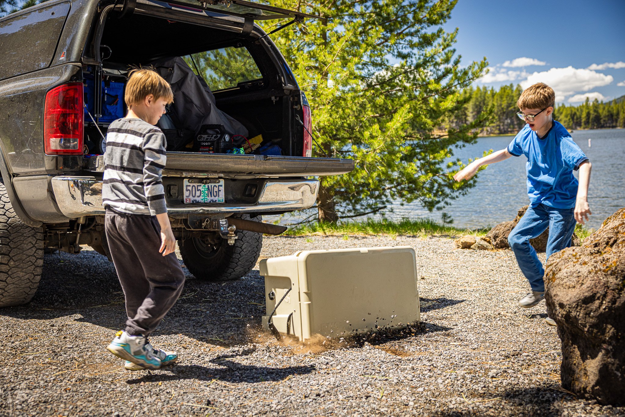 Two boys are playing outside near a lake with rocks on the ground and a large tree in the background. One boy in a striped shirt is standing while the other in a blue shirt is running or jumping. An open truck with its bed filled with camping gear is