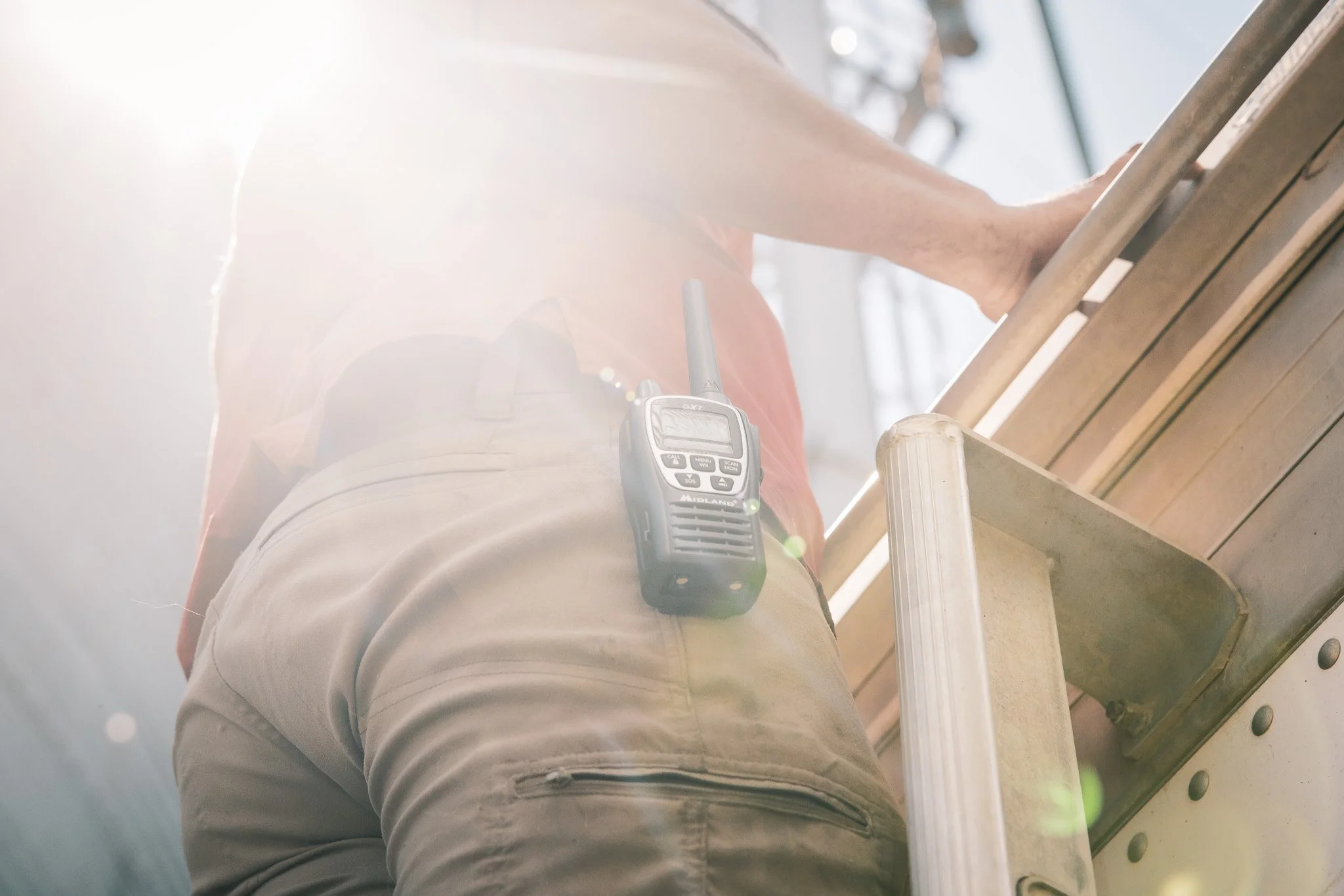 Person climbing a ladder with a walkie-talkie clipped to their pants, sunlight shining from behind.