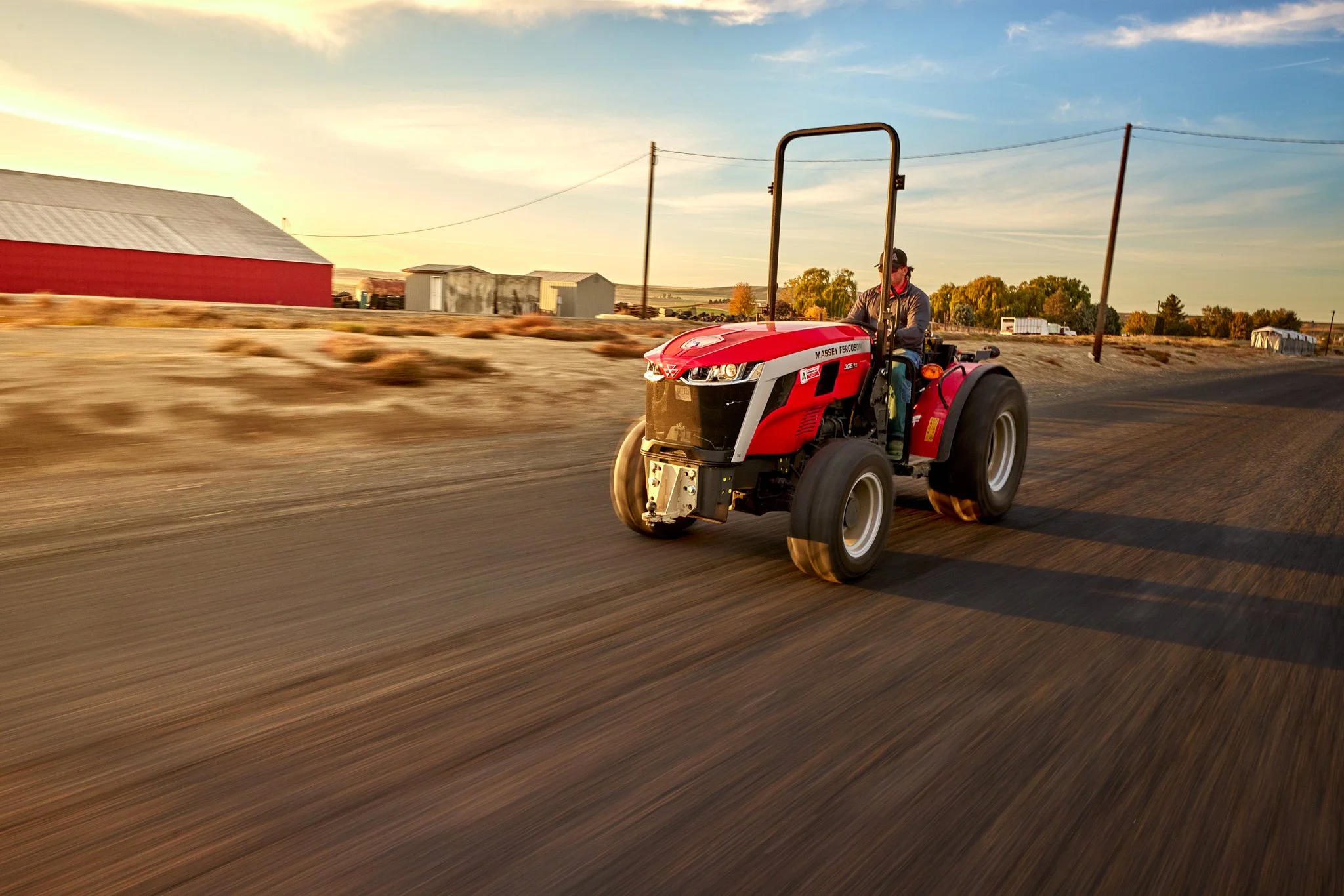 A man driving a red Massey Ferguson tractor on a dirt road at sunset, with farm buildings and power lines in the background.
