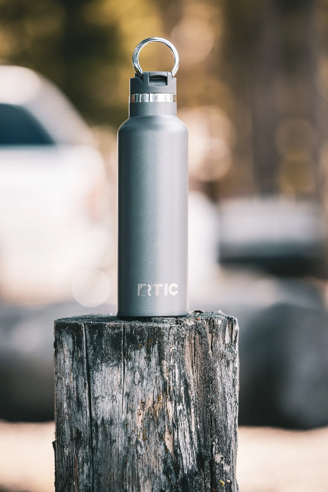 Gray RTIC water bottle with a black lid and silver loop handle on a rustic wooden post. Background is blurred outdoor scene.