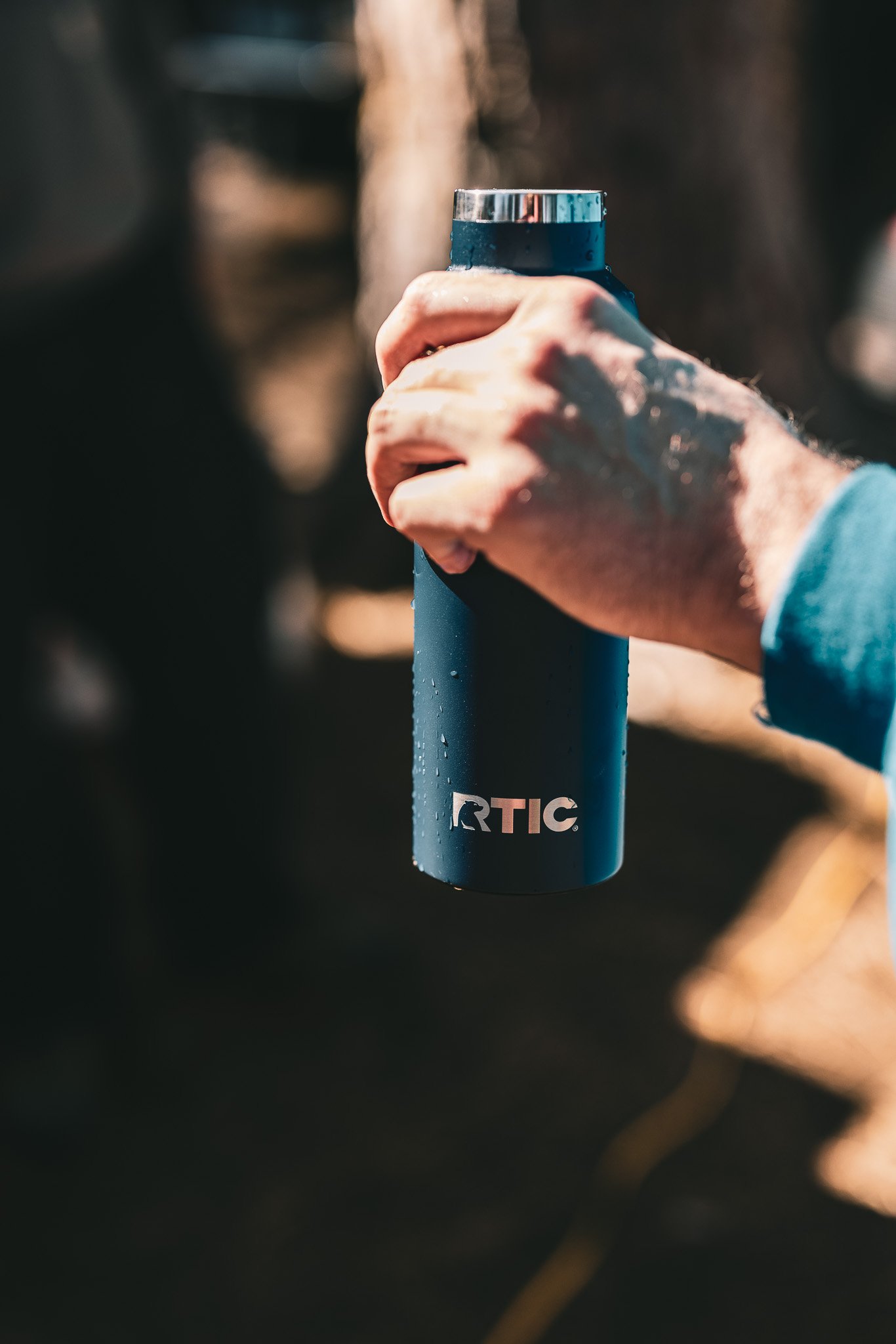 A person holding a blue RTIC tumbler outdoors. The tumbler has water droplets on it, and the person is wearing a blue jacket.