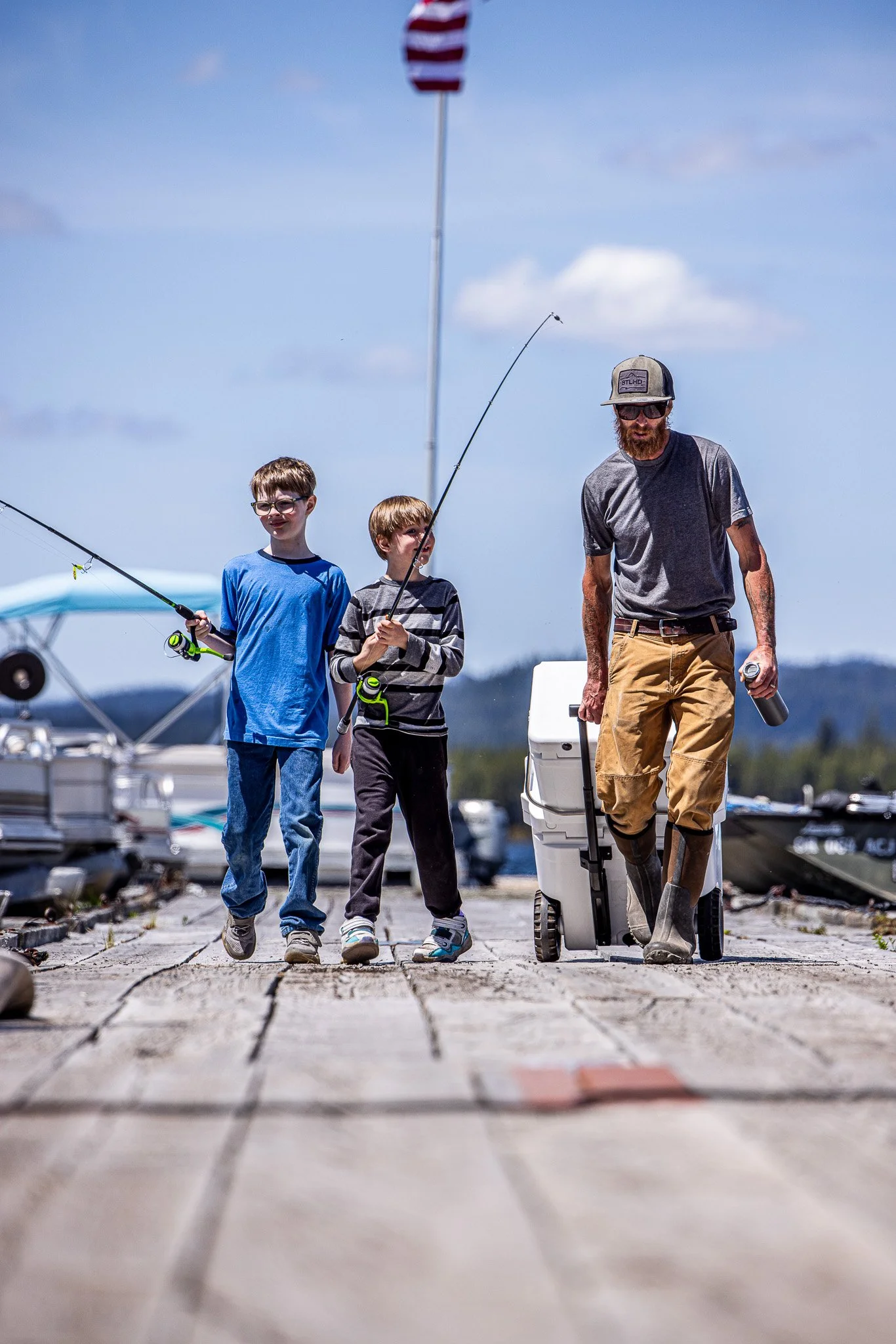 An adult man and two boys walk along a dock with boats. The children hold fishing poles, and an American flag is visible in the background.