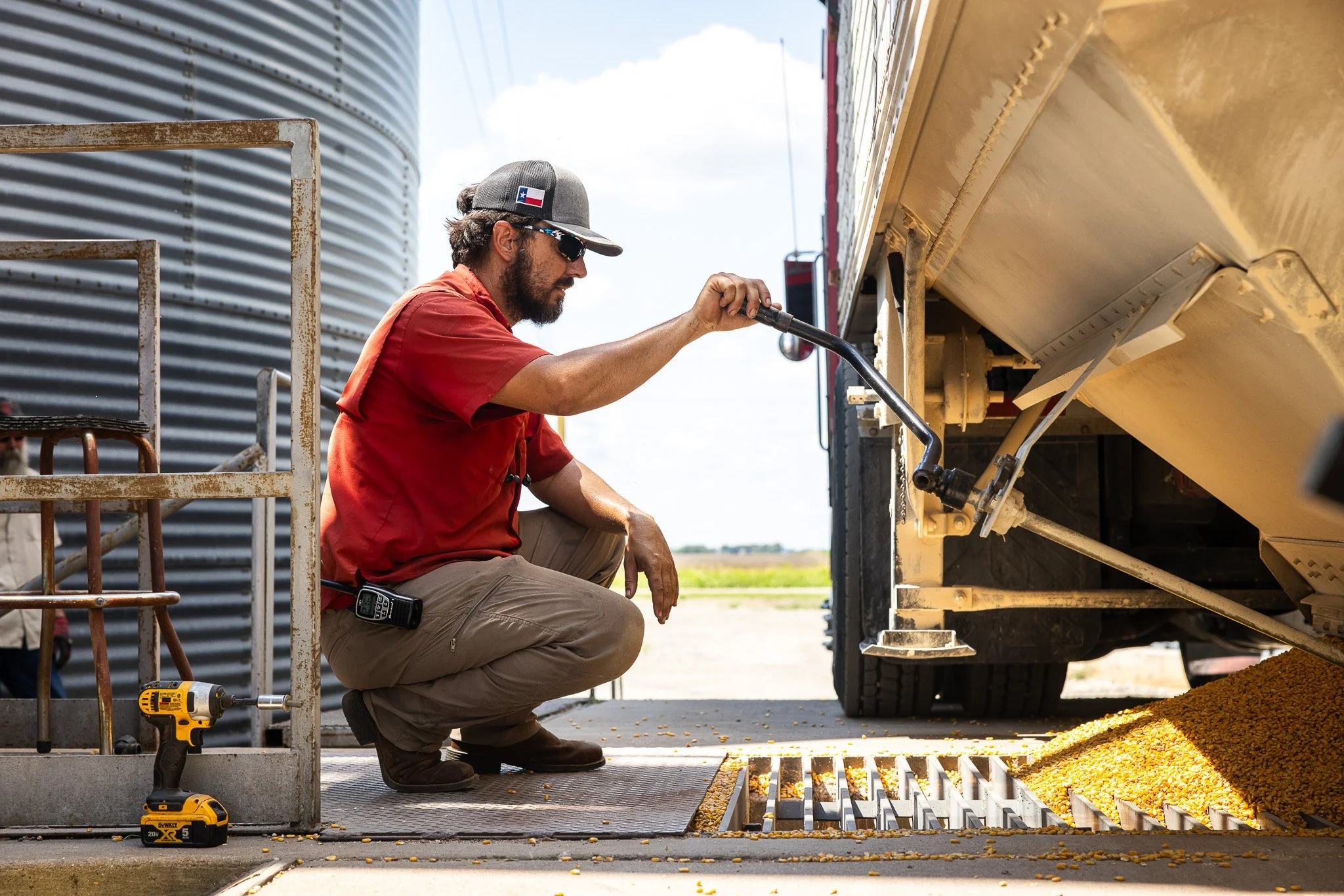A man in a red shirt, beige pants, and sunglasses is kneeling next to a truck, fueling it with corn from an open grain tank. He is holding a nozzle to the tank, with a small yellow cordless drill on the ground nearby.