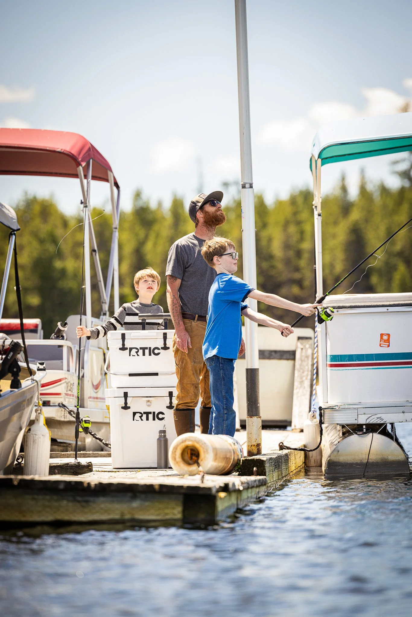 Three males fishing from a dock on a lake with boats in the background during daytime.