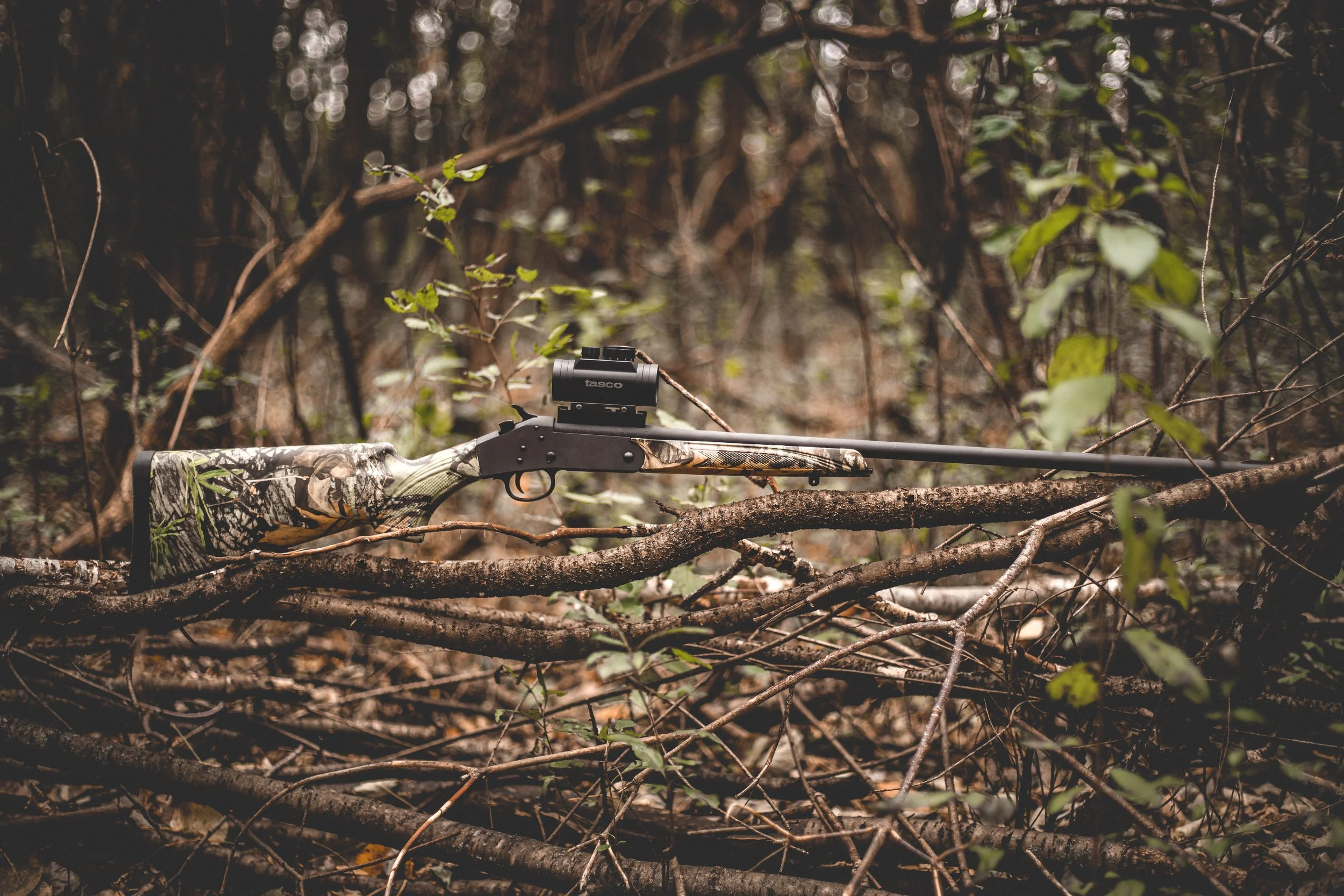 A camouflage deer hunting rifle with a scope resting on fallen branches in a dense forest.