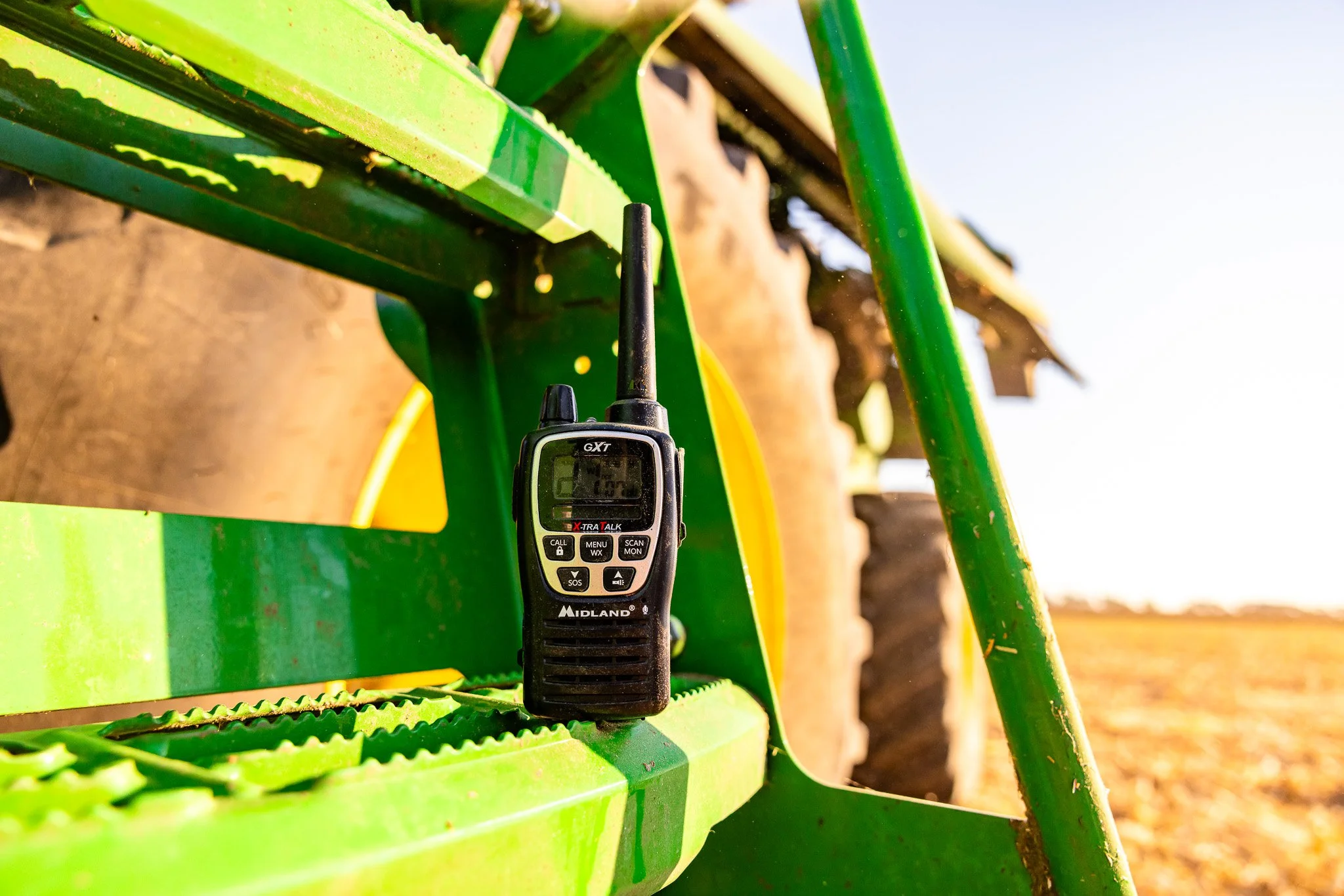 A handheld radio transmitter placed on a green tractor's metal surface in a farm field during daylight.