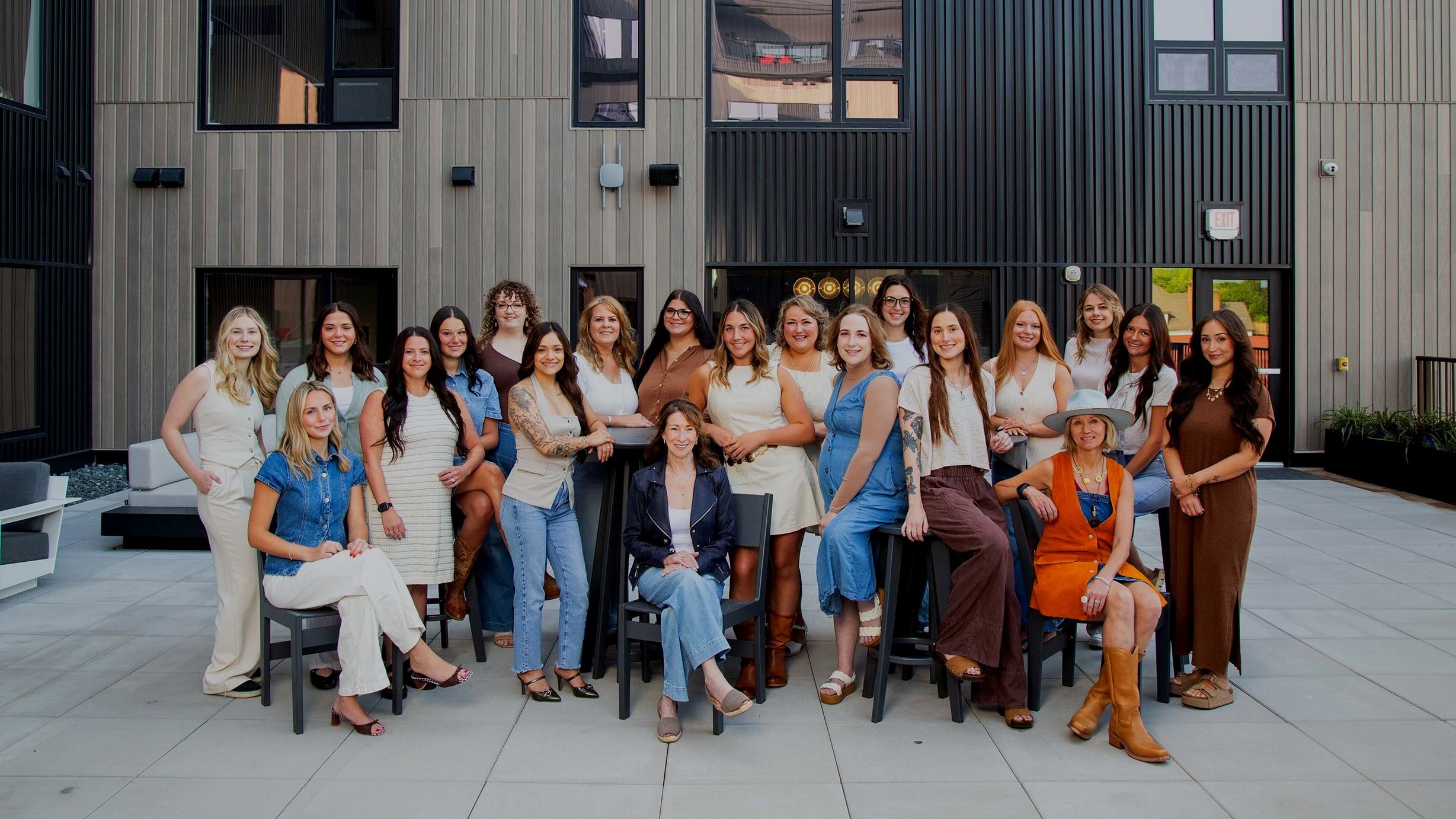 Group of women posing outdoors on a patio in front of a modern building with large windows.