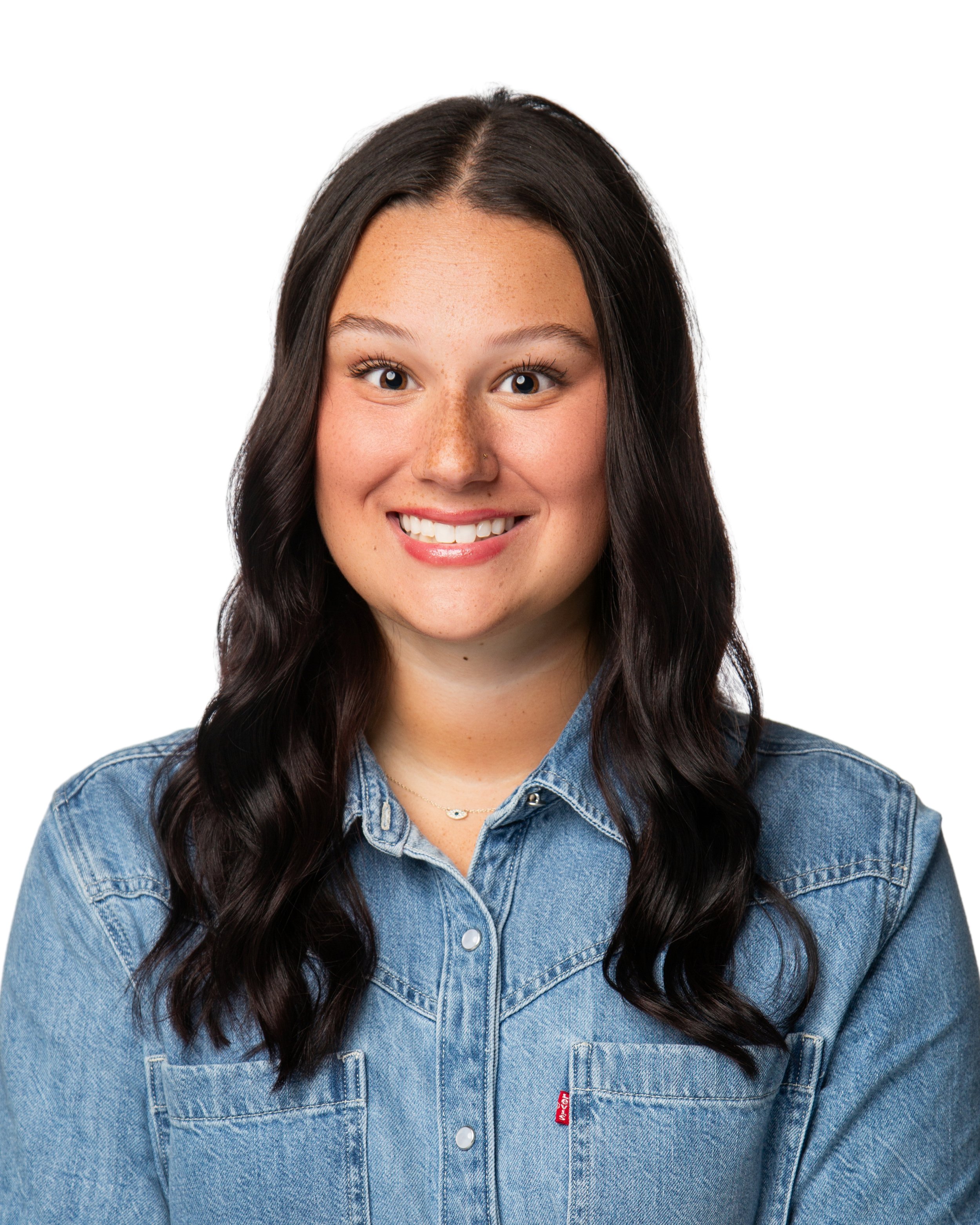 Close-up of a smiling young woman with long dark wavy hair, wearing a blue denim jacket, against a plain white background.