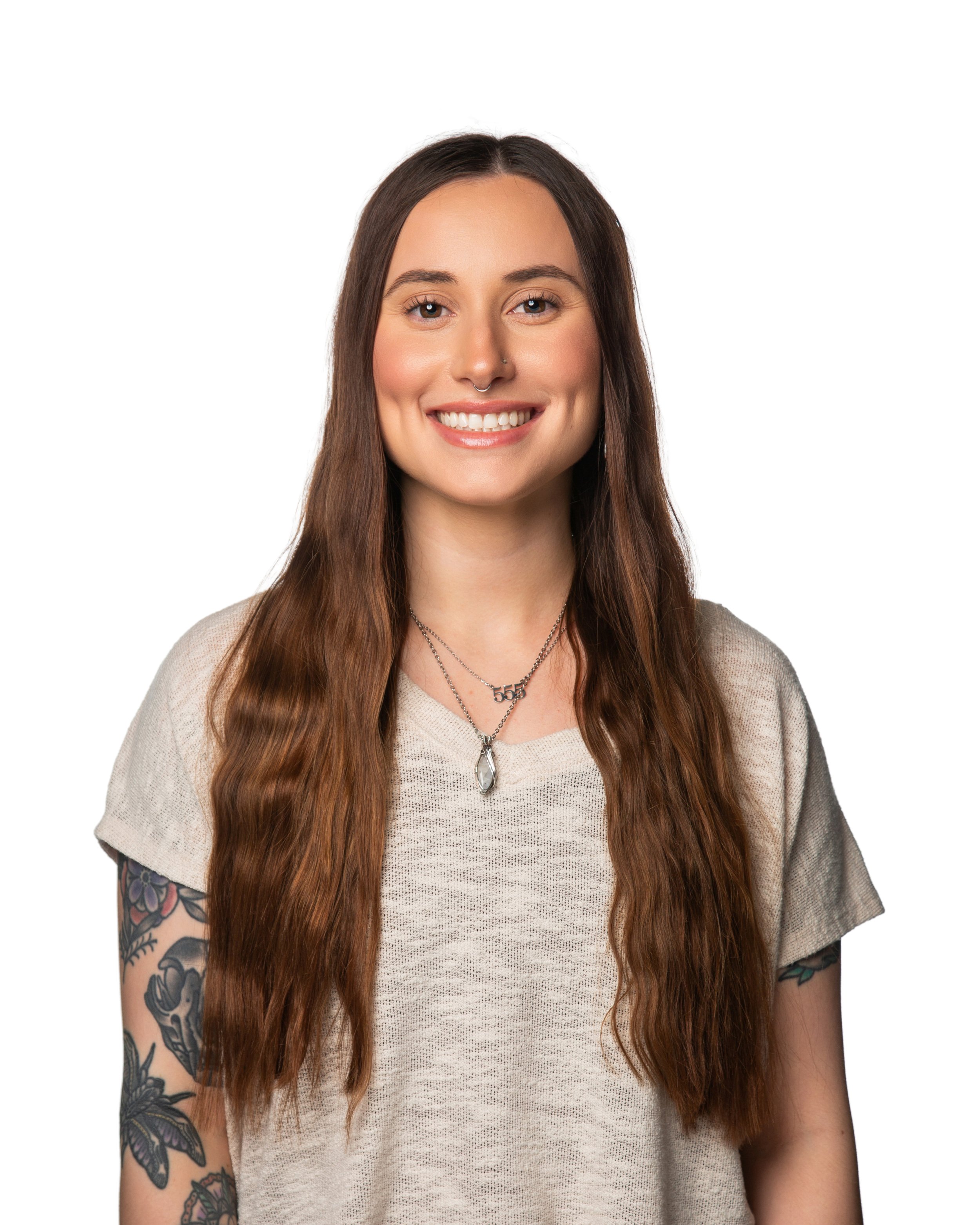 A young woman with long brown hair, tattoos on her right arm, smiling with straight white teeth, wearing a beige sweater and layered necklaces, against a plain white background.