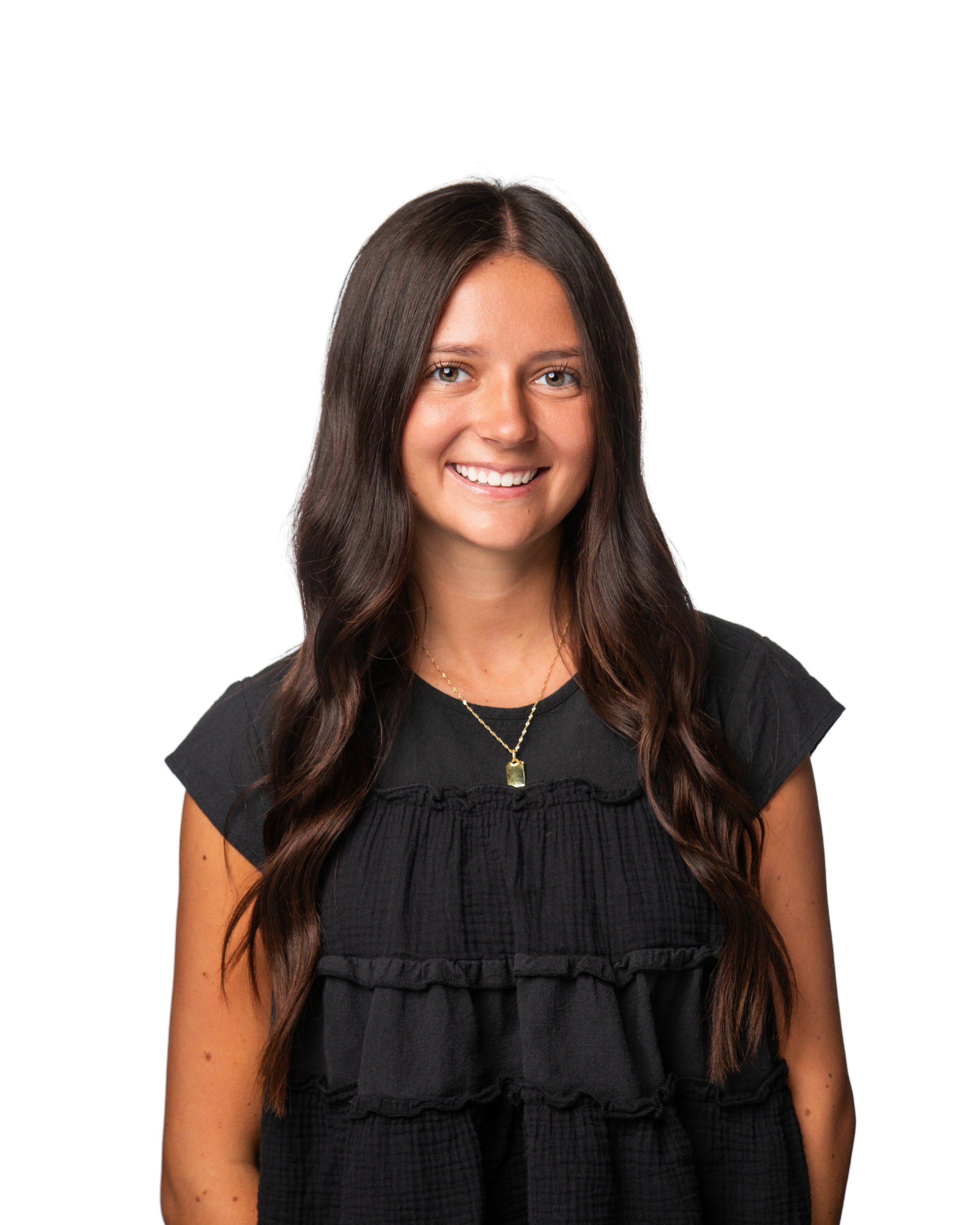 Close-up of a smiling woman with long wavy dark brown hair wearing a black top with ruffled details and a gold necklace with a rectangular pendant, against a plain white background.