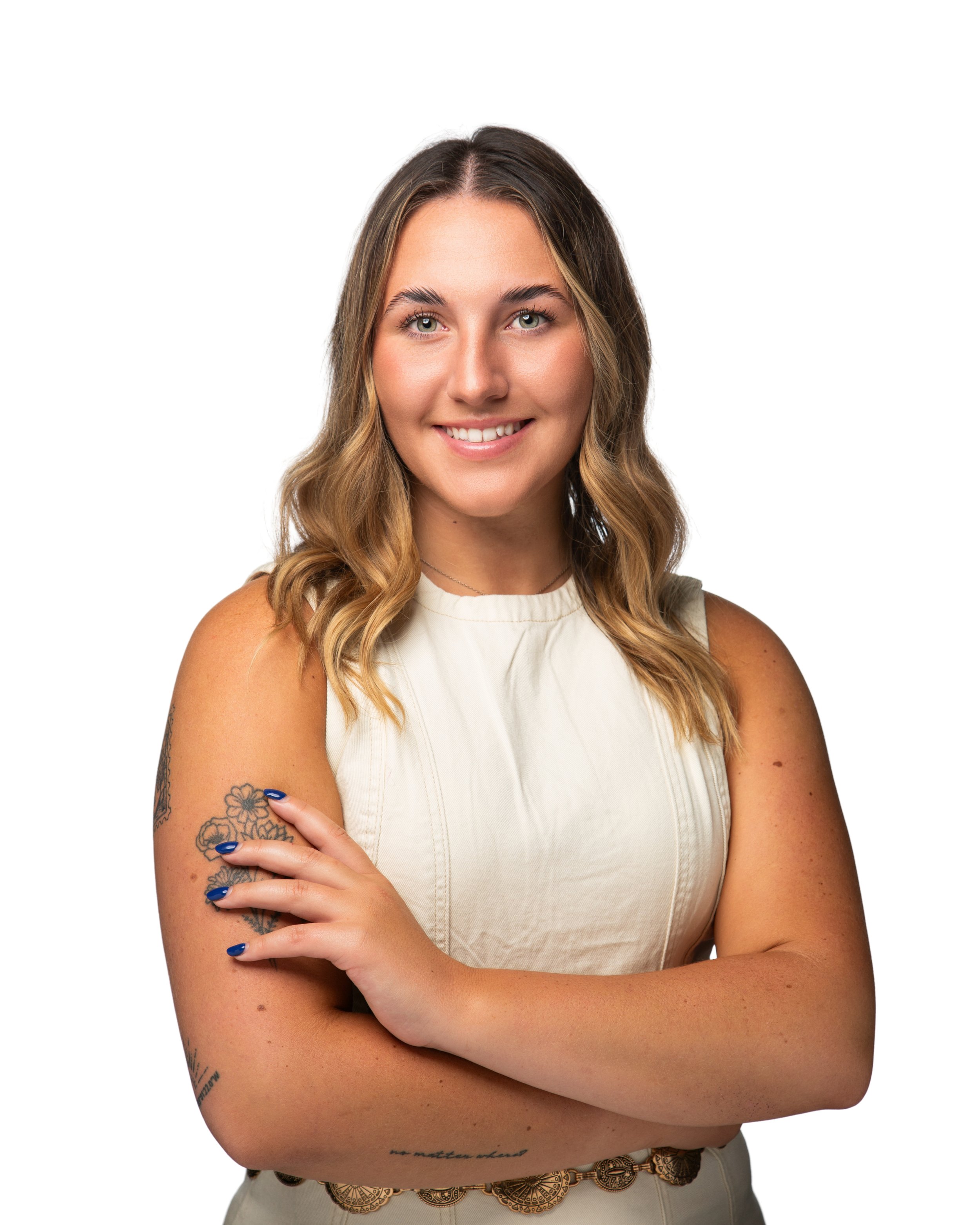 A woman with long wavy hair, light skin, and tattoos on her arm and waist, smiling with arms crossed, wearing a beige sleeveless top and a decorative belt, against a plain white background.