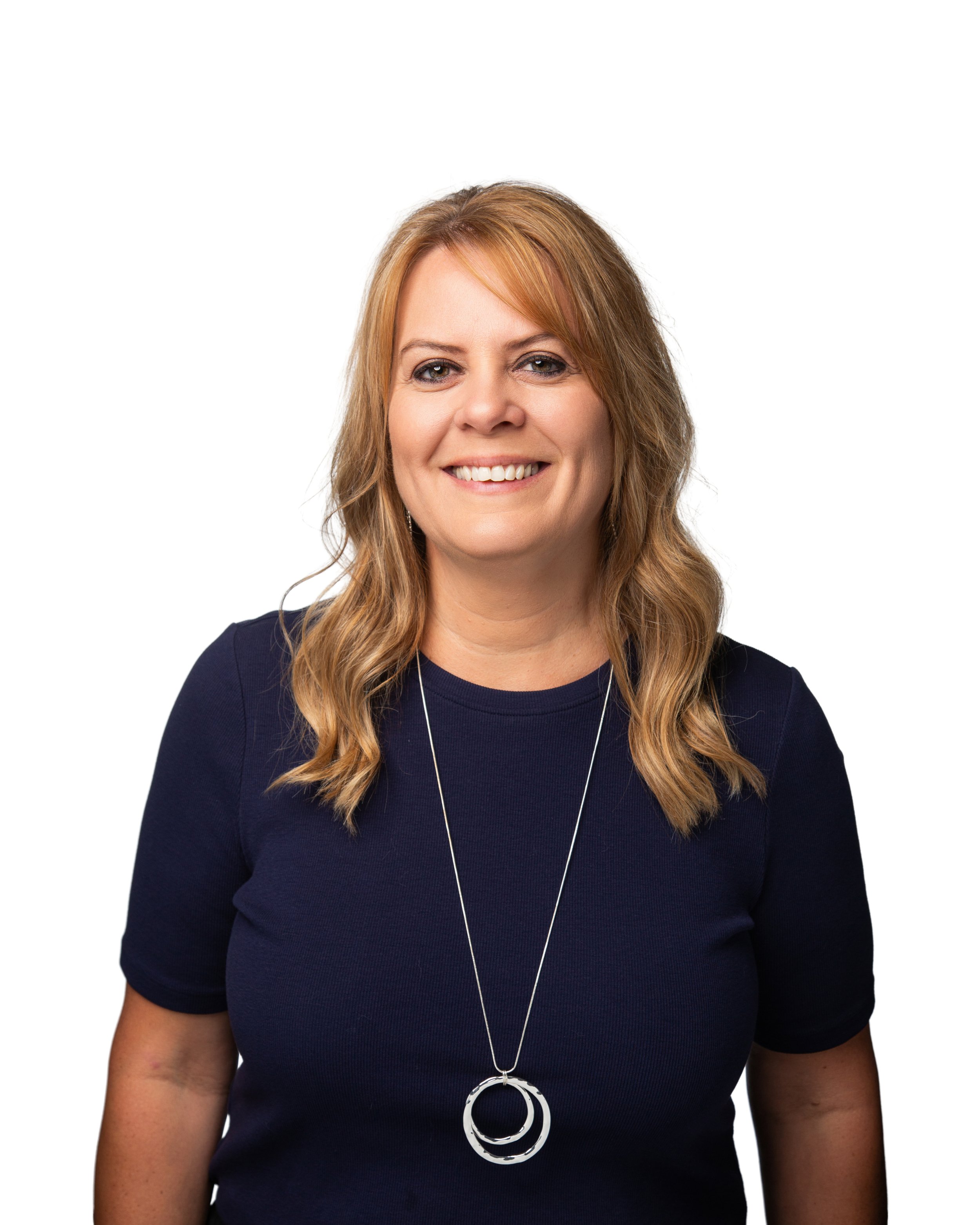 Close-up of a smiling woman with wavy light brown hair wearing a dark navy shirt and a long silver necklace with multiple rings.