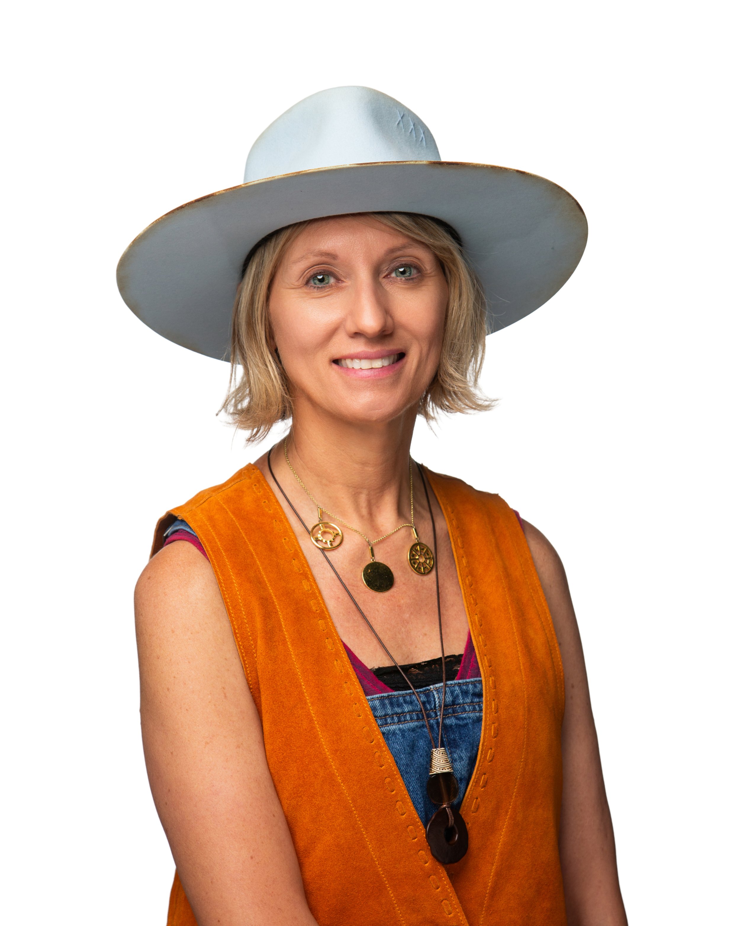 A woman with blonde hair wearing a large white hat, an orange vest, layered necklaces, and colorful clothing, smiling with her arms crossed.