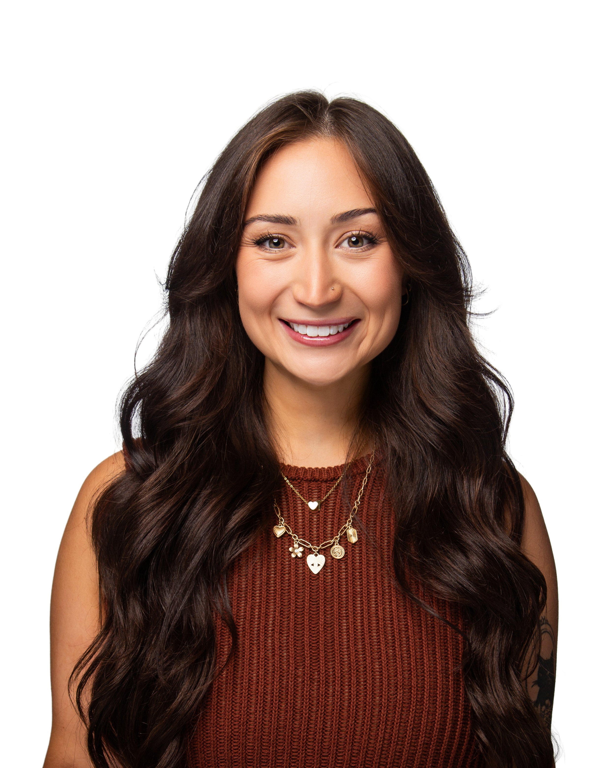 Close-up of a smiling woman with long, wavy brown hair, wearing layered gold necklaces and a sleeveless, textured rust-colored top, against a white background.
