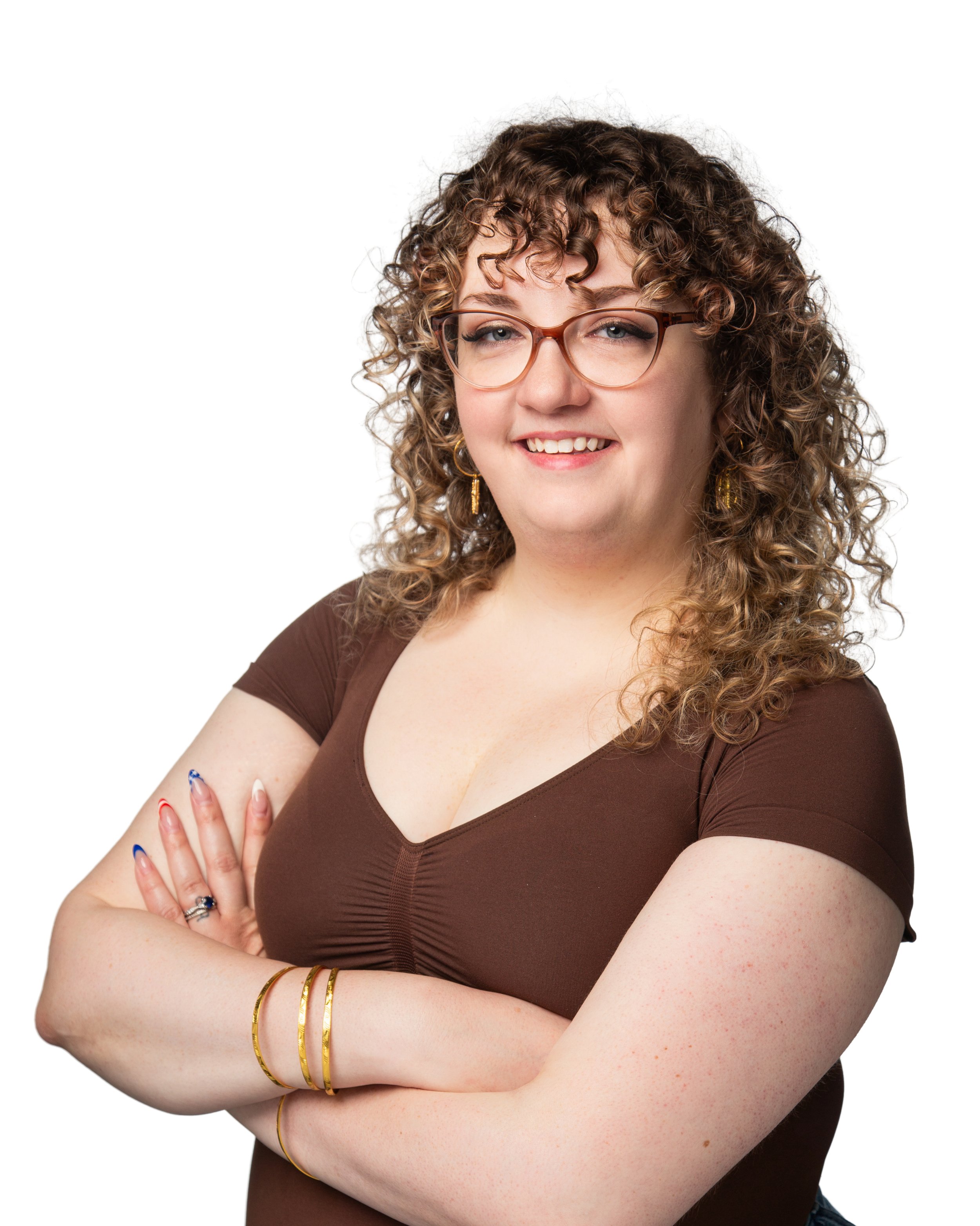 A woman with curly brown hair, wearing glasses, a brown top, and gold jewelry, standing with arms crossed and smiling at the camera.