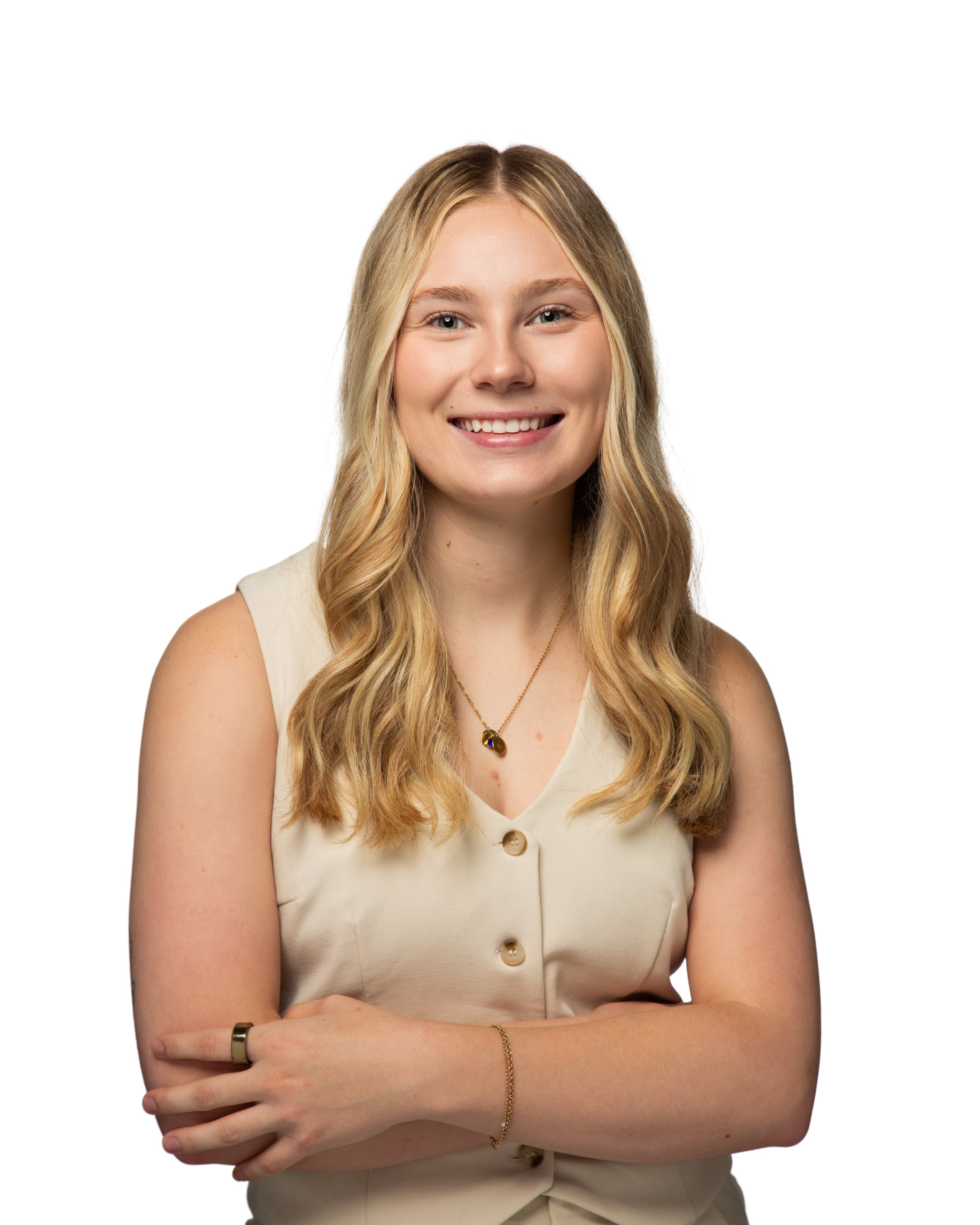 Close-up of a woman with long blonde hair smiling, wearing a beige sleeveless button-up top, with gold jewelry including a necklace and bracelet, against a white background.