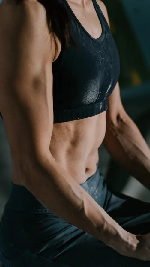 Close-up of a woman in athletic wear showing her toned abs and arms, sitting in a fitness environment.