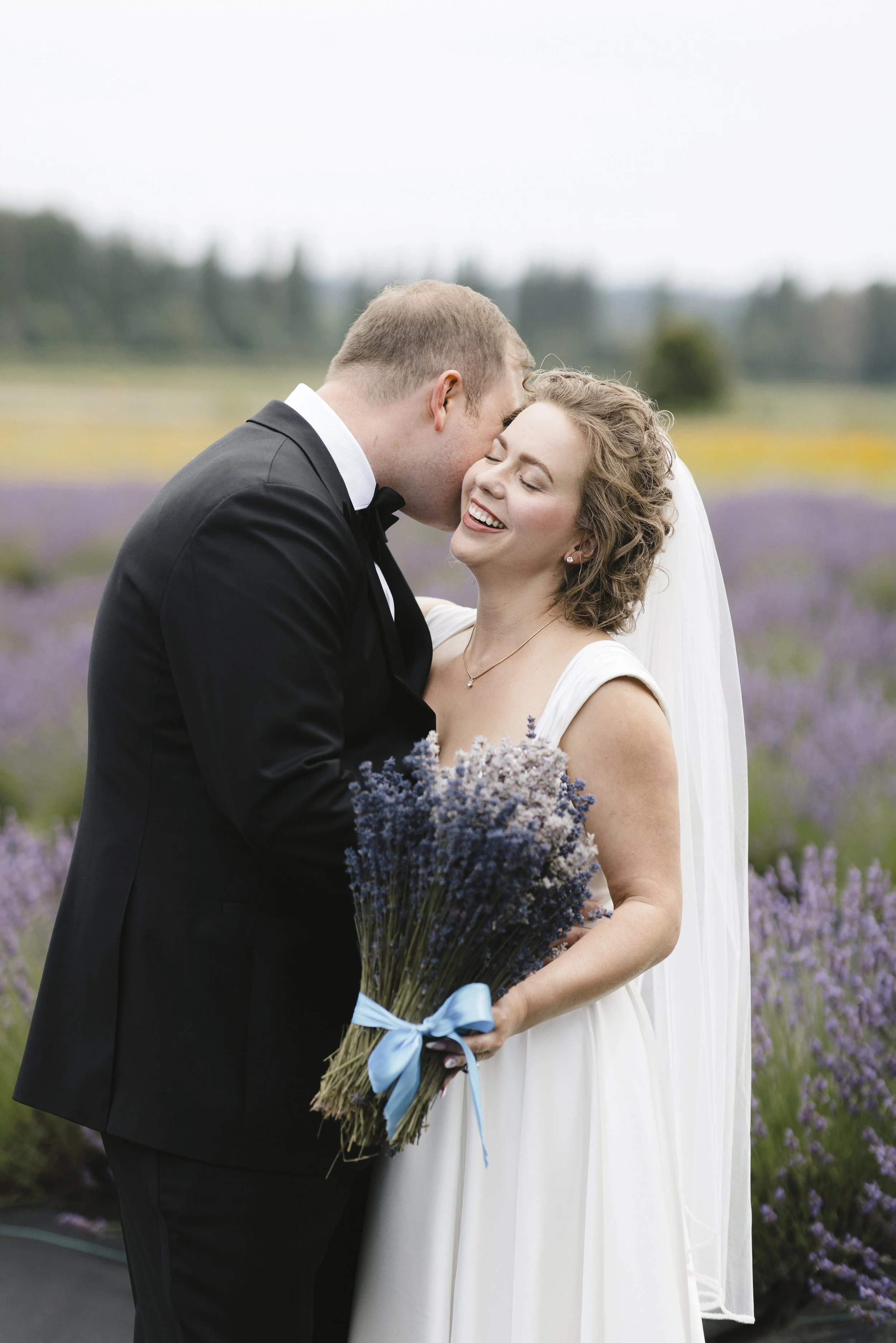 A newlywed couple sharing a kiss outdoors in a lavender field, with the bride holding a bouquet of lavender tied with a blue ribbon.