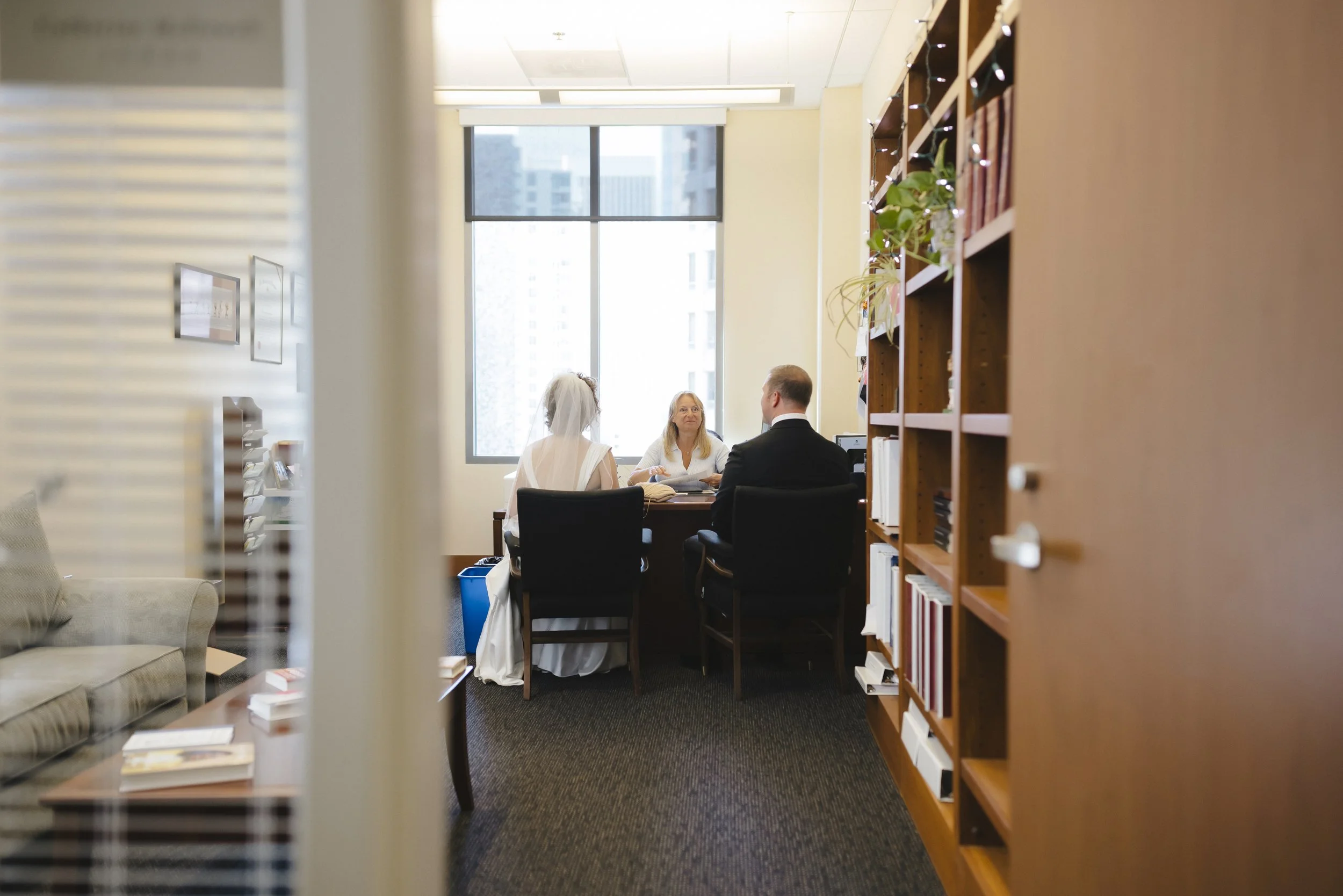 A wedding ceremony takes place in an office with a bride and groom facing an officiant, seated at a desk near a window, with bookshelves on the right side.