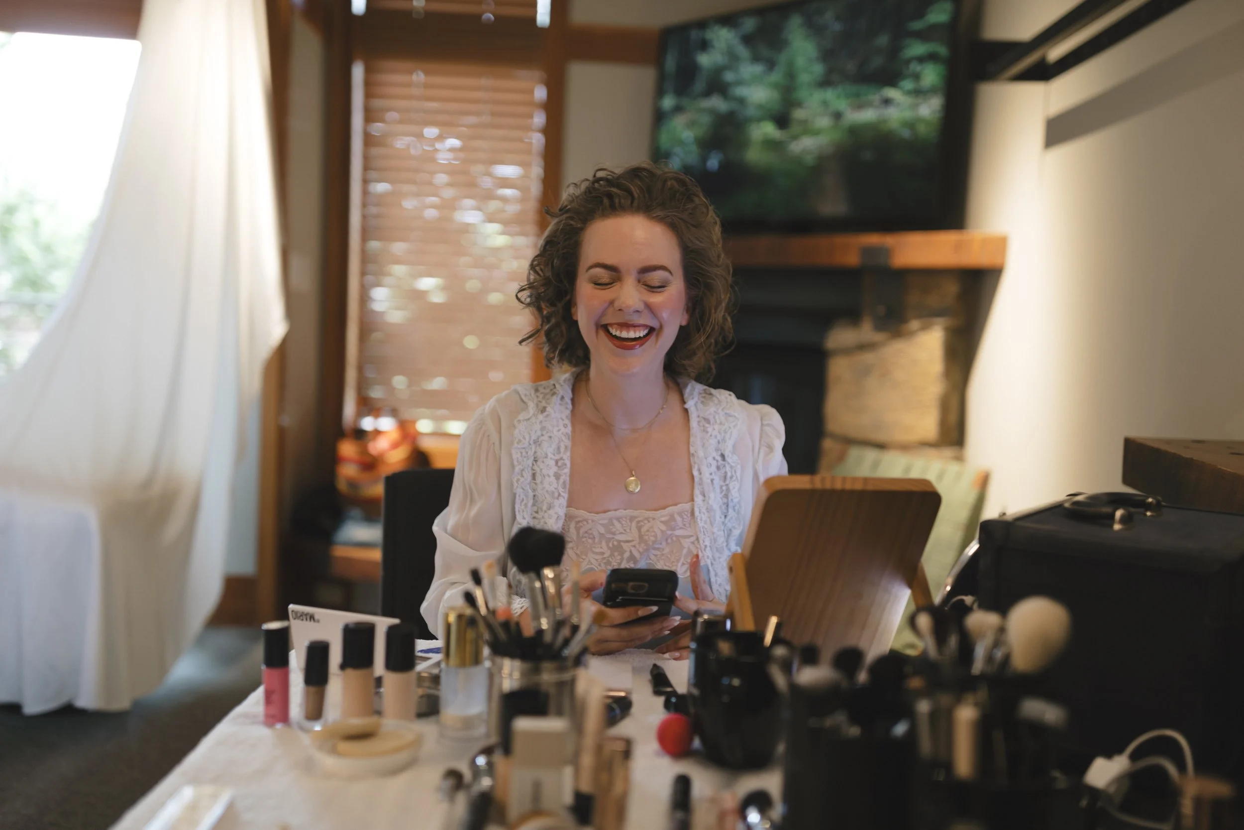 A woman with curly hair smiling while looking at her phone in a room with makeup products on a table.