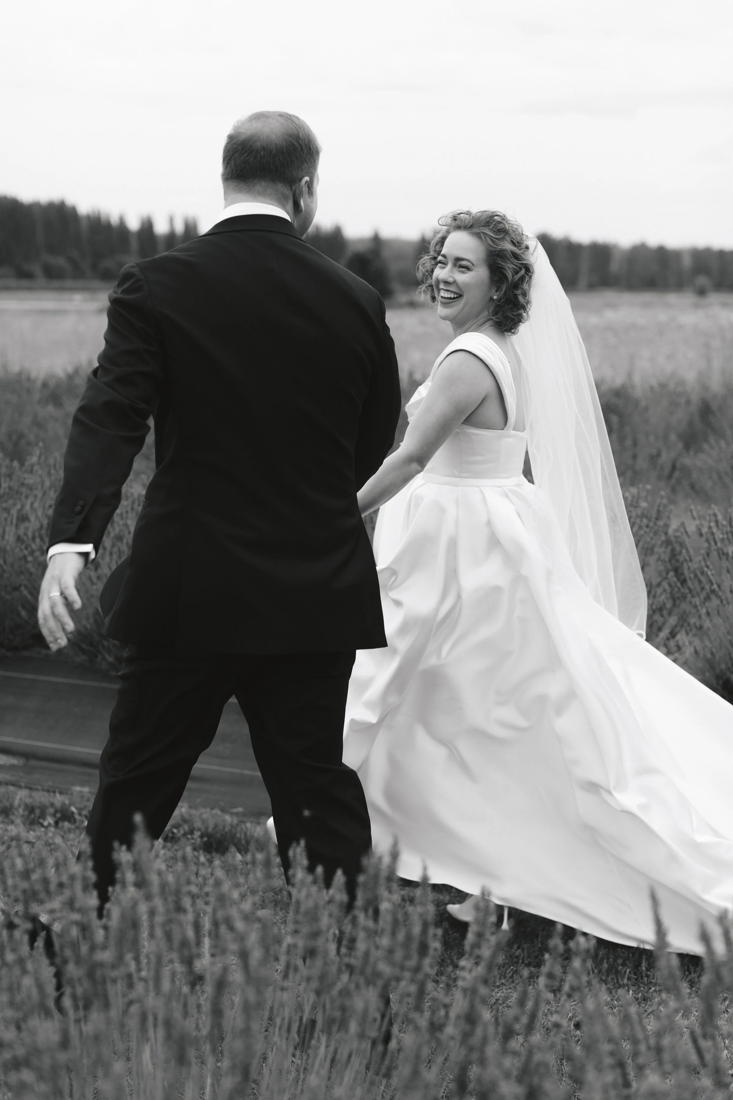 Black and white photo of a bride and groom outdoors, holding hands, with the bride smiling and looking back at the camera, in a field.