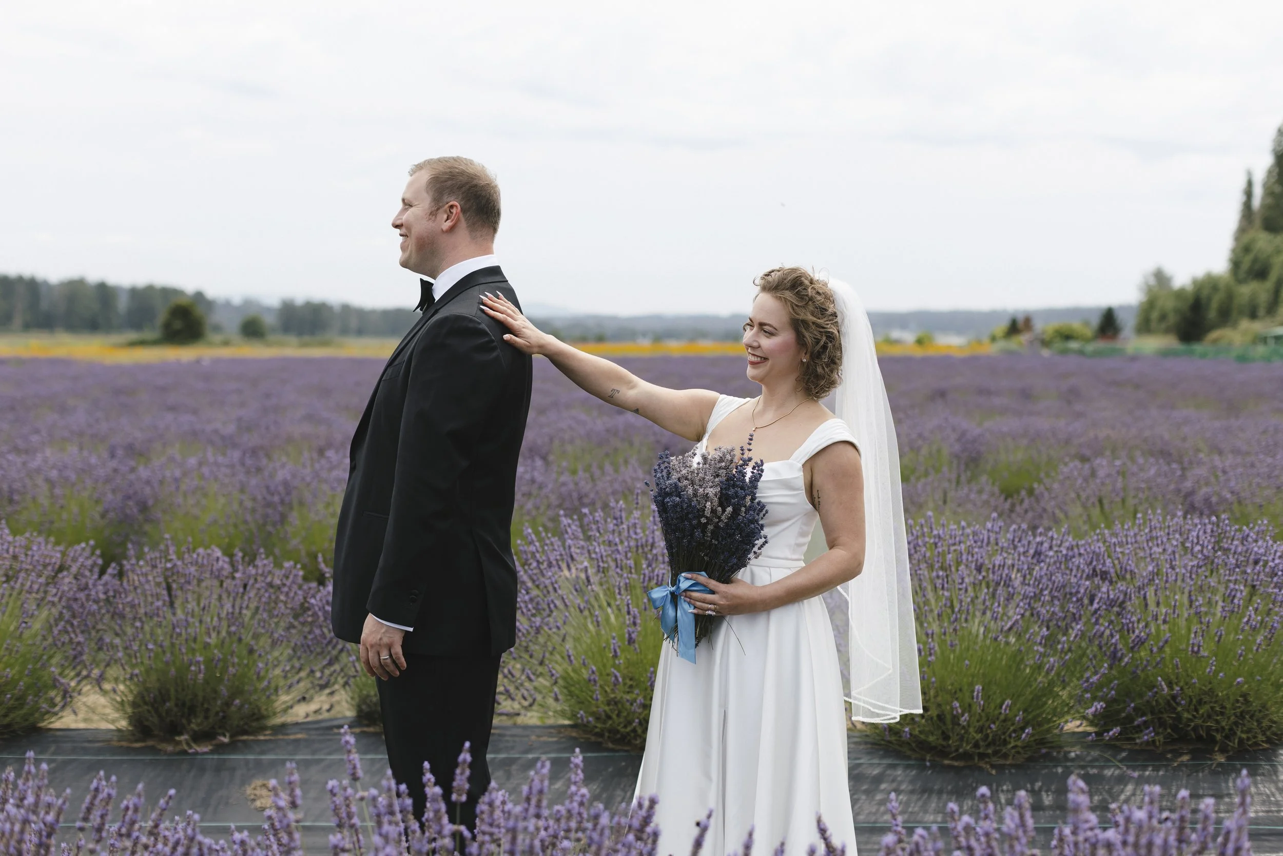 A bride touches a groom's shoulder in a lavender field during a wedding photoshoot, with the bride holding a bouquet of lavender flowers and smiling.