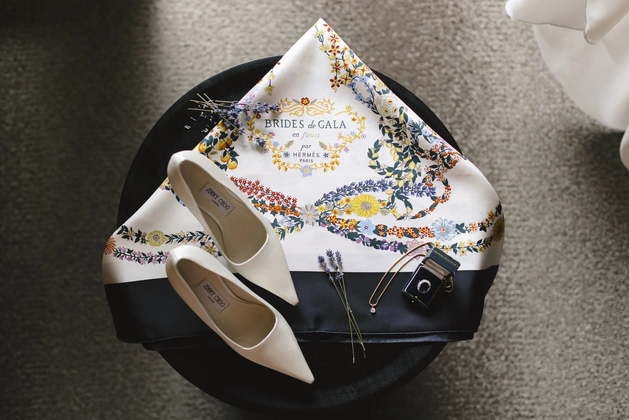 A black round table with a floral scarf, white high-heeled shoes, lavender sprigs, a gold necklace with a pink pendant, and a miniature black camera on top, with a textured gray carpet underneath.