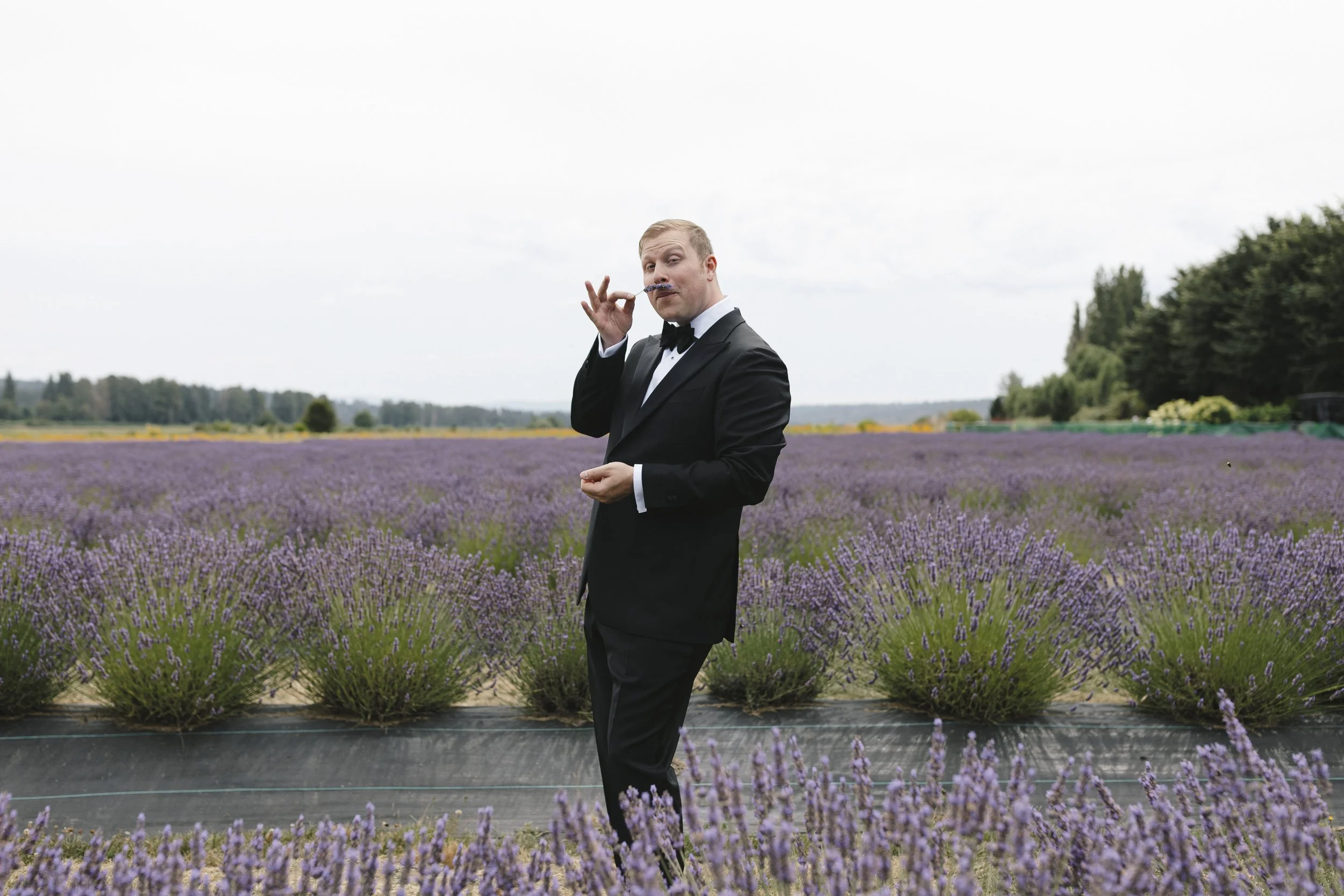 A man in a tuxedo standing in a lavender field with purple flowers and green trees in the background.